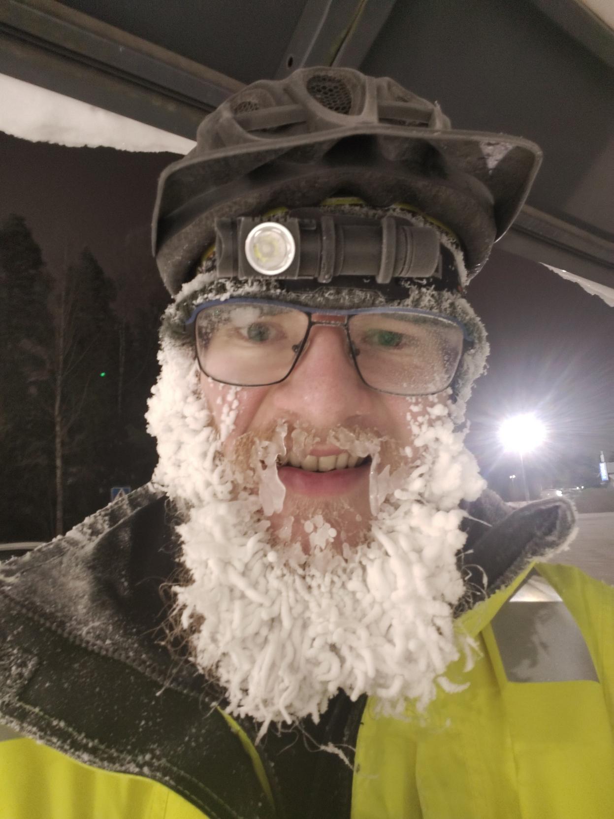 Kuurapartainen pyöräilijä. Parta on valkoista tykkylunta ja viiksistä roikkuu ainakin kaksi senttiä pitkät jääpuikot, joiden takaa pilkottaa jotain hymyn tapaista. Taustalla aamun pimeys ja kirkas keinovalo. Yllä näkyy pyöräkatos.