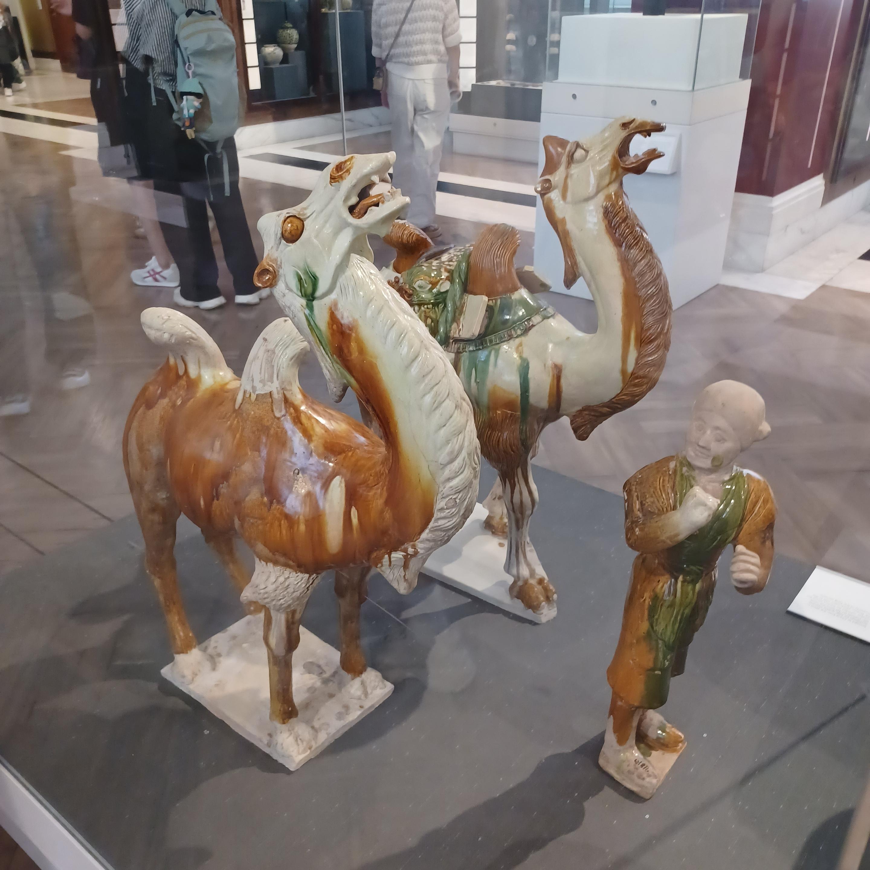 Valokuva kolmesta Tang-dynastian aikaisesta lasitetusta keramiikkapatsaasta. Kaksi suurta patsasta esittää kaksikyttyräisiä kameleita, jotka nostavat pitkiä kaulojaan ja näyttävät mylvivän. Kamelipatsaat on maalattu punaruskealla ja vihreällä lasitteella. Toisella kameleista on satula. Kolmas patsas, pieni ihmishahmo, seisoo kameleiden edessä ja näyttää vetävän niitä näkymättömällä köydellä. Tämäkin patsas on maalattu ruskealla ja vihreällä lasitteella. Sillä on päällään pitkä kauhtana. Patsaat ovat suuren vitriinin sisällä ja niiden takana näkyy museovierailijoita.