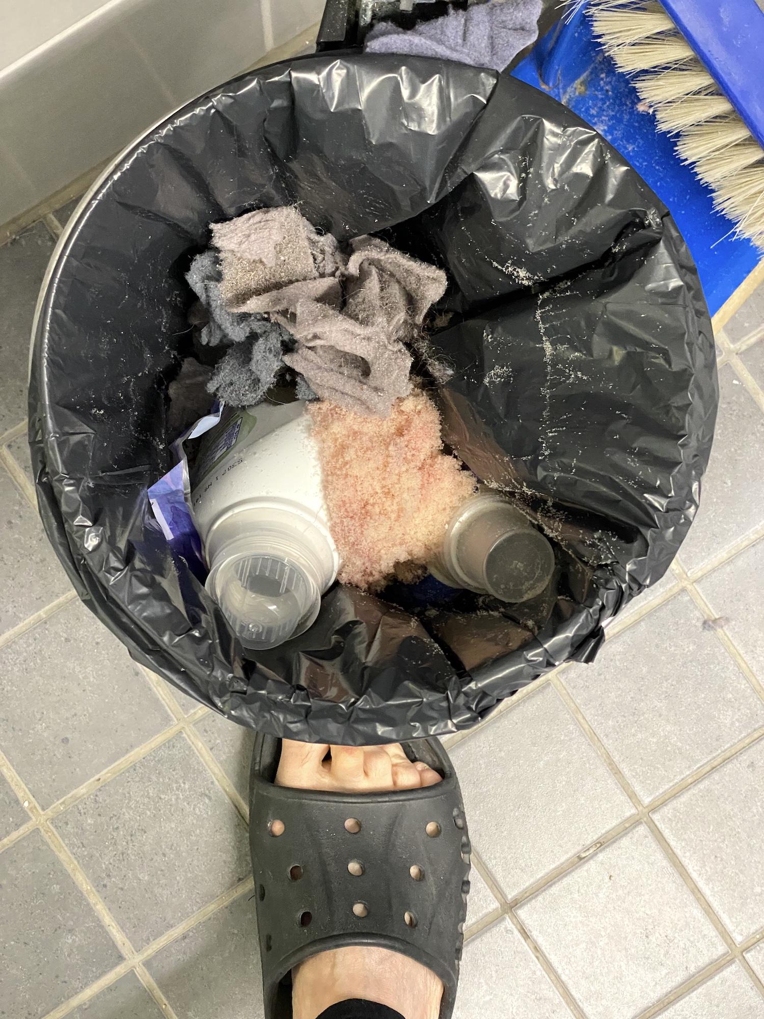 Ylhäältä kuvattu pieni roskapönttö, jossa on nukkapaakkujen ja muun lian seassa kaksi muovista pesuainepulloa. Pöntön alla kantta polkimella auki pitävä sandaaliin pukeutunut jalka.