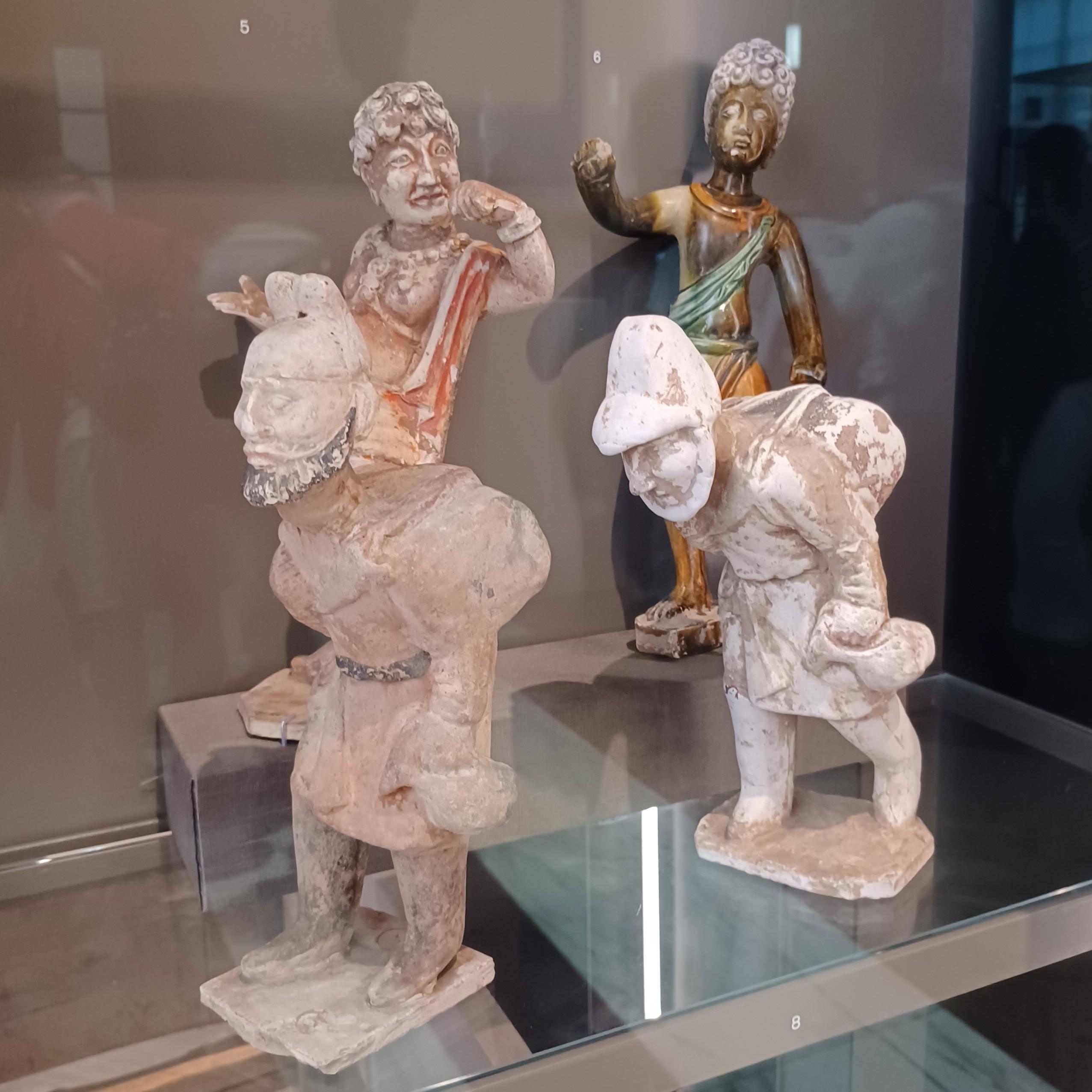 Valokuva neljästä keramiikkapatsaasta. Etualalla on kaksi patsasta, jotka esittävät hiukan kumarassa käveleviä miehiä, jotka kantavat selässään taakkoja ja käsissään juomakannuja. Miehillä on päähineet ja parrat, polviin ulottuvat vyötetyt kauhtanat sekä saappaat. Takana olevat patsaat esittävät parrattomia miehiä, joilla on lyhyt, kihara tukka. Toinen on maalattu tummaihoiseksi tummanruskealla lasitteella. Molemmilla on kaulakääty ja paljaan ylävartalon päällä toisen olan yli kiedottu punainen tai vihreä kangasvaate. Hahmot kohottavat käsiään eleisiin, ikään kuin pitäisivät puheita.