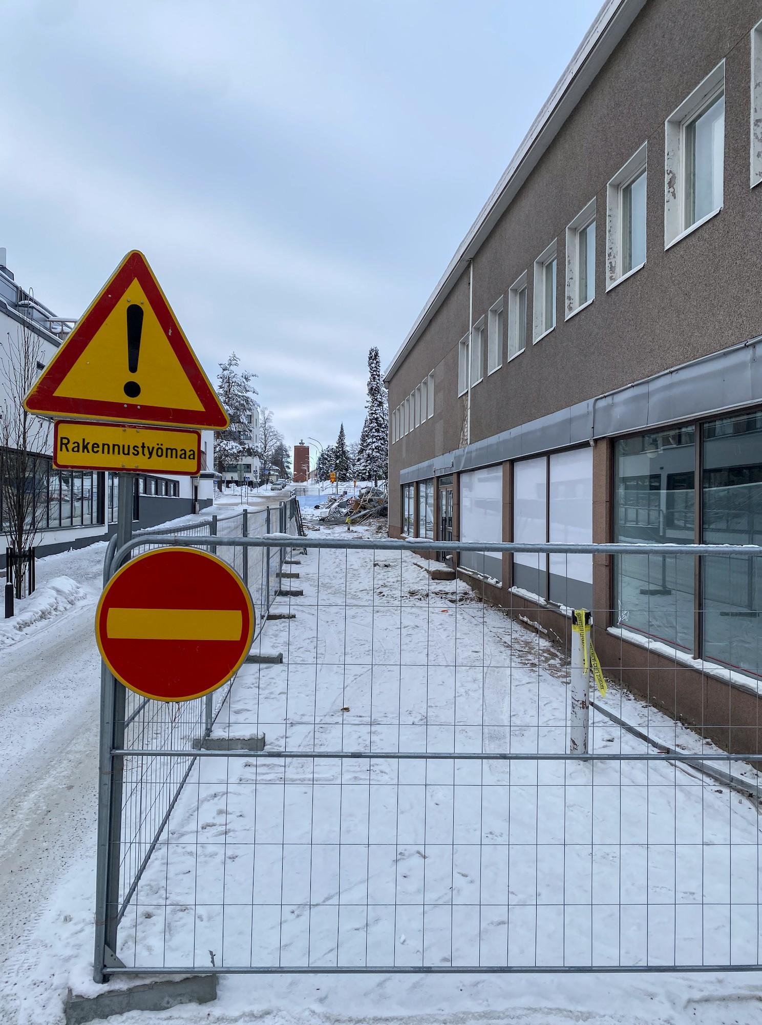 Näkymä lumista katua pitkin loivaan ylämäkeen. Kadun oikealla puolella on kaksikerroksinen ruskeaksi rapattu liikerakennus, jonka katukerroksessa on näyteikkunoita ja toisessa kerroksessa neliömäisten ikkunoiden rivi. Talo ja jalkakäytävä on ympäröity työmaa-aidalla. Aidan vasemmassa kulmassa on liikennemerkkejä: muu vaara ja lisäkilpi "Rakennustyömaa" ja sen alla kielletty ajosuunta. Korttelin vastakkaisesta päästä noin puoliväliin taloa on purettu. Tien varrella ylempänä on lumisia kuusia ja kaukana tien päässä punatiilinen torni.