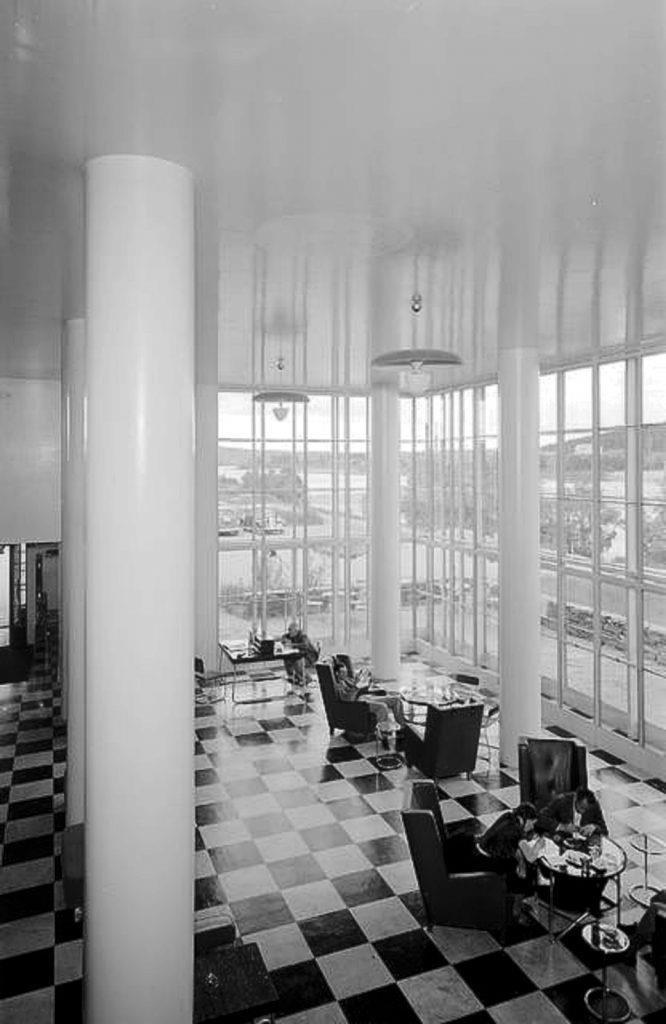 Mustavalkoinen valokuva korkeasta funkkistyylisestä hotelliaulasta, jossa on valkoisia pylyväitä, šakkiruutulattia ja ikkunat lattiasta kattoon.