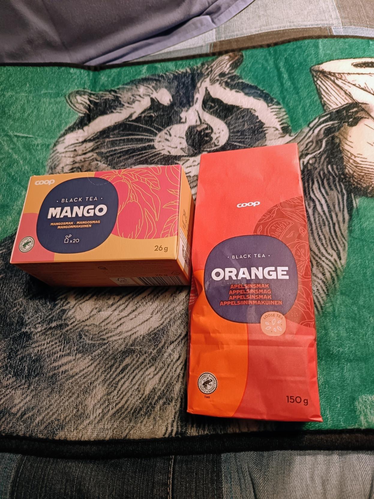 pikkumatto, jossa pesukarhu ojentaa vessapaperia.
maton päällä vasemmalla mangotee ja oikealla appelsiiniteepussukka.