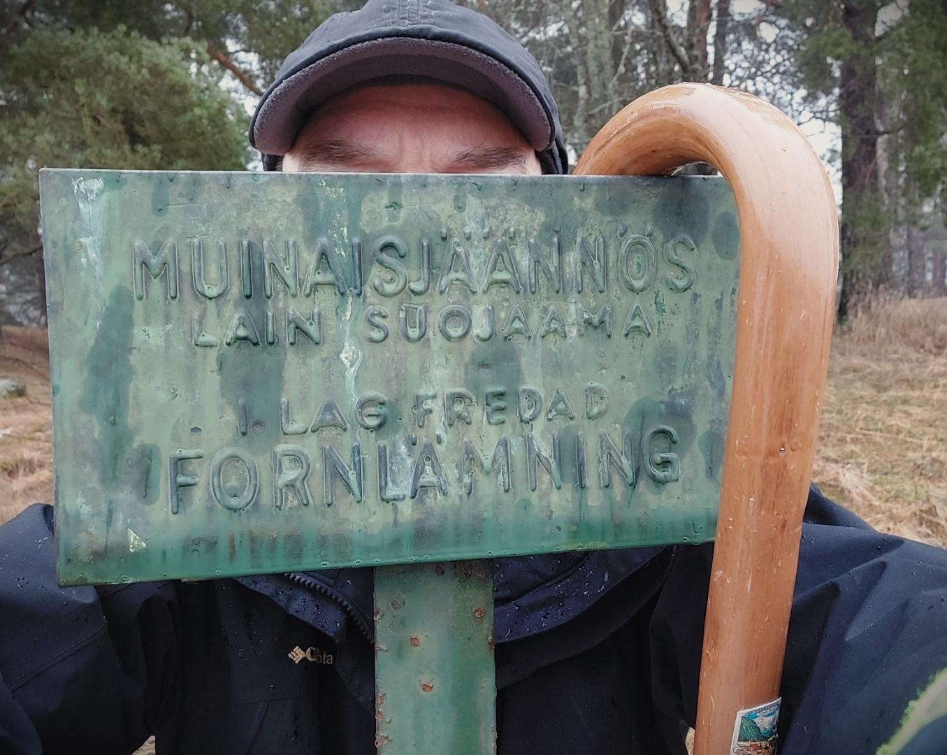 Onnellinen kuvaaja piiloutunut Komoistenkumpujen Muinaisjäännös-kyltin taakse Mallnitzilaisine kävelykeppeineen. Vihreässä kyltissä vielä enemmän ajan patinaa kuin kulkijan varusteissa ja habituksessa. Kyltti julistaa: "Lain suojaama".