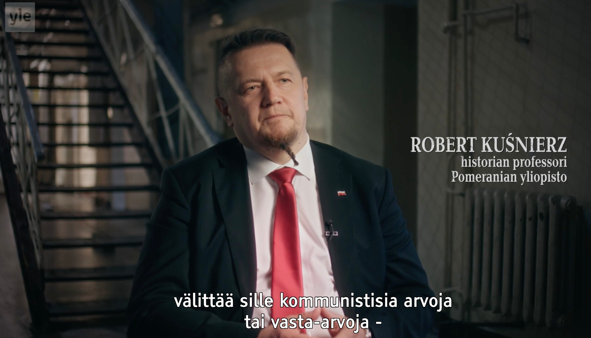 Pysäytyskuva YLE Areenasta. Käytävällä portaikon edessä istuu kasvot kameraan päin keski-ikäinen pukumies, parta pienellä palmikolla, valkoinen paita, punainen solmio ja rintapielessä Puolan lippu -pinssi. Vieressä teksti: ROBERT KUŚNIERZ, historian professori, Pomeranian yliopisto.