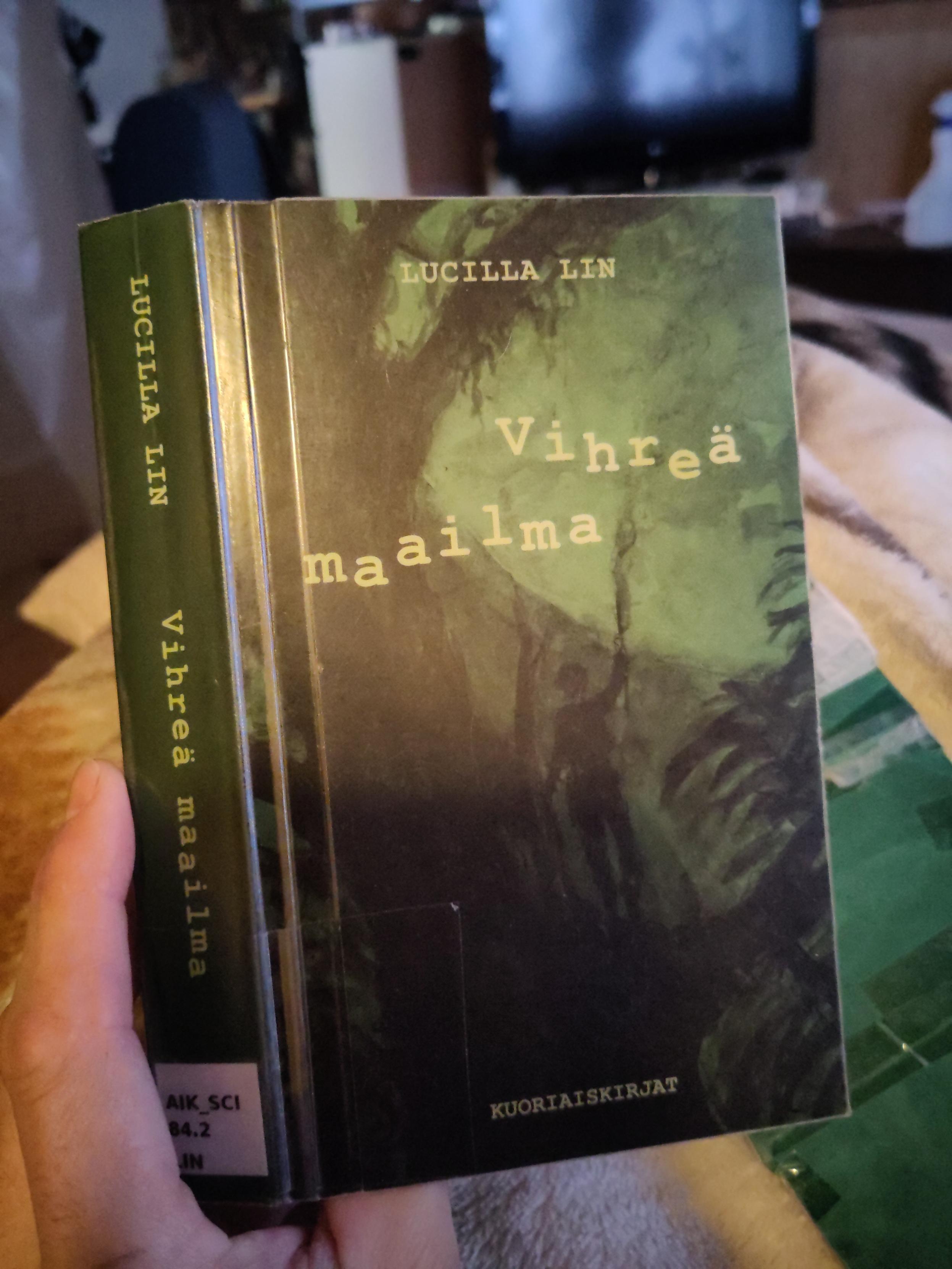 Kädessä vihreä kirja, Lucilla Lin Vihreä maailma.
Vihreällä taustalla tumma puu ja ihmishahmo