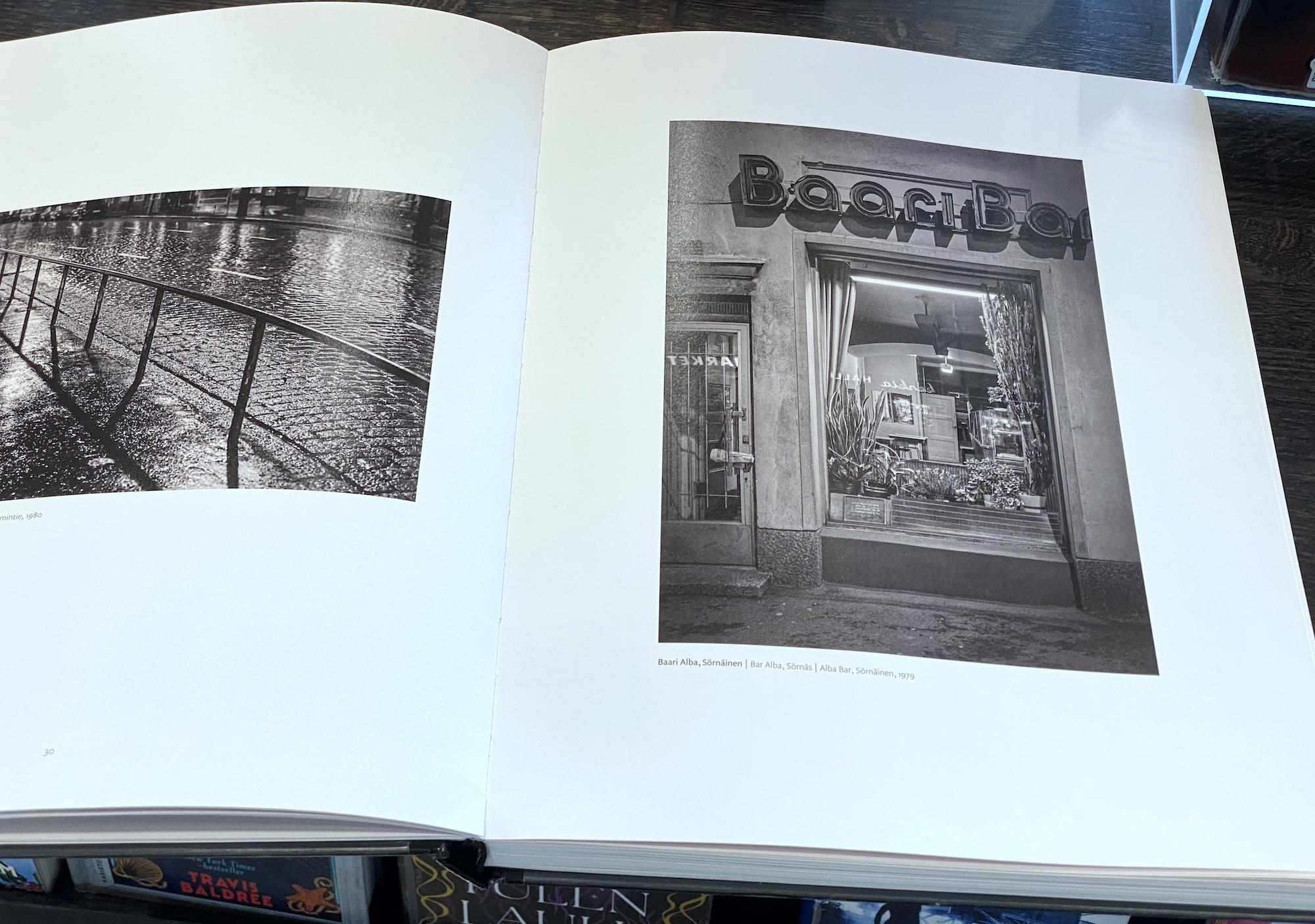 Valokuvakirjan mustavalkoinen aukeama. Vasemmalla sivulla sateinen mukulakivikatu, jossa kosteus kimaltelee. Oikealla baarin julkisivu: ovi, ikkuna jossa viherkasveja ja yllä valokirjaimet "BaariBar", joissa kuitenkaan ei pala valot.