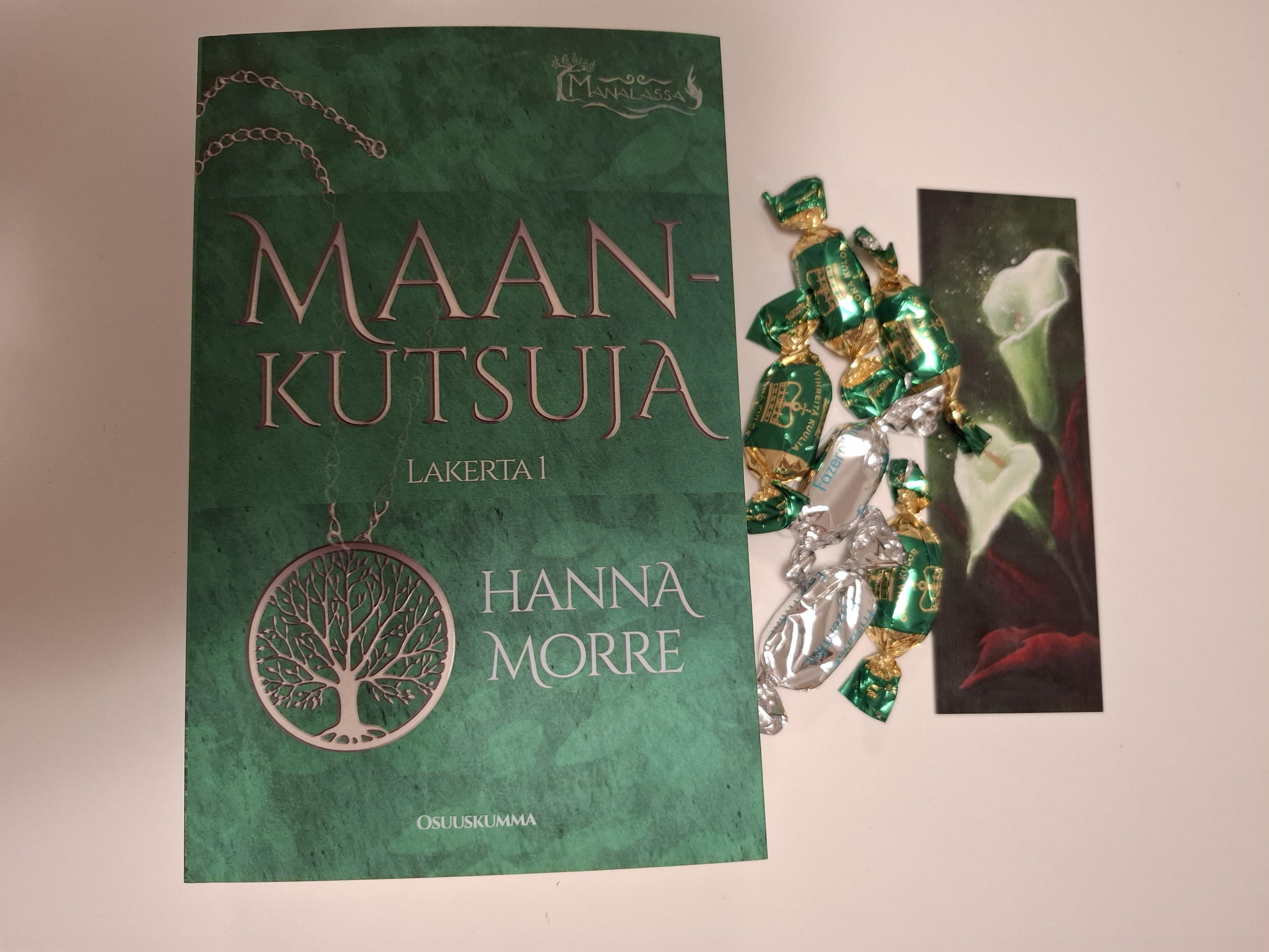 Hanna Morren Maankutsuja-kirjan kansi, jossa on vihreällä taustalla puuta esittävä riipus. Vieressä karkkeja ja kirjamerkki.
