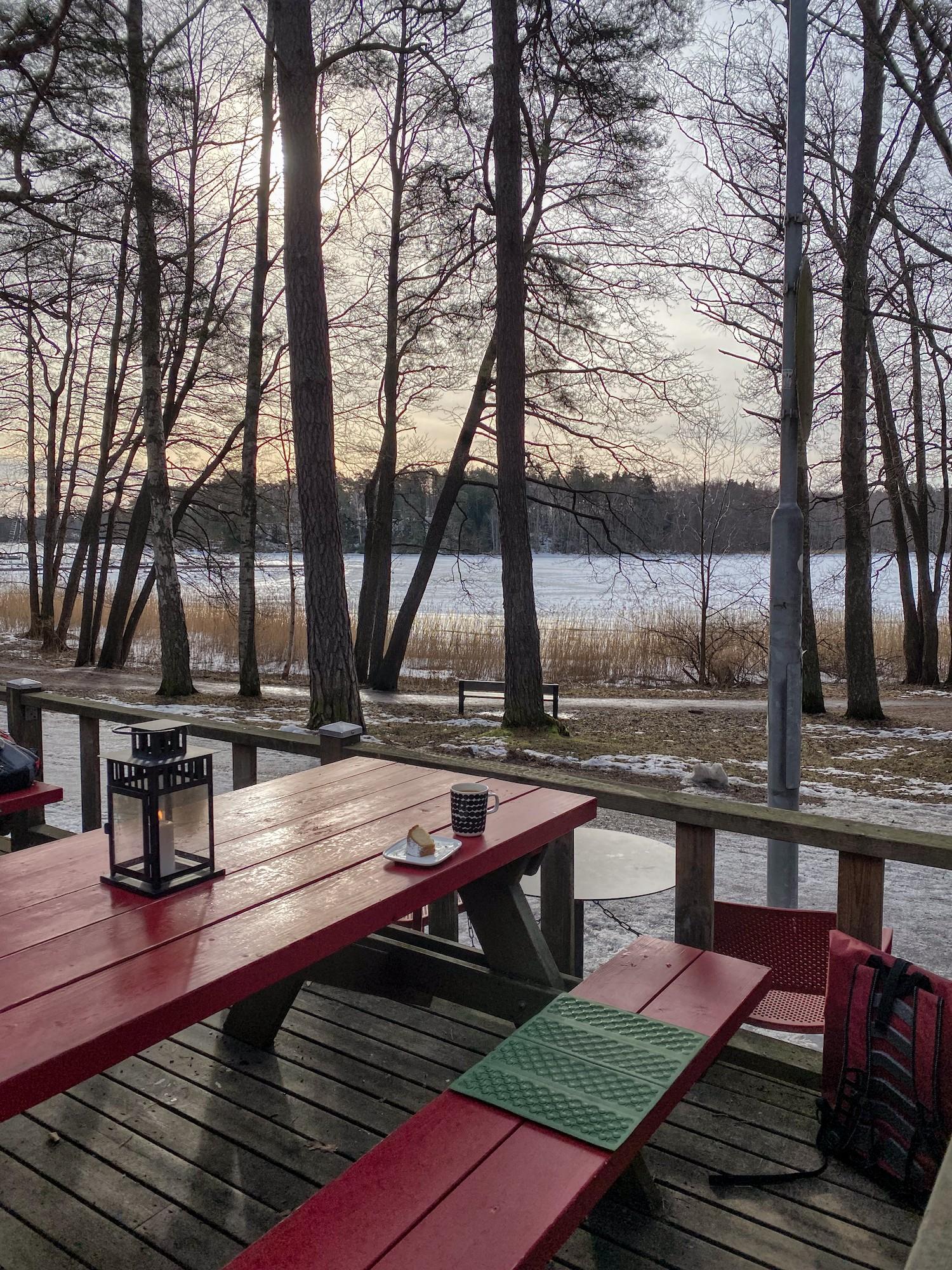 Kahvilan jykevä puinen ulkopöytä ja penkki. Pöydällä on kynttilälyhty, kahvikuppi ja kakkulautanen, jolla olevasta kuivakakkupalasta on haukattu kerran. Penkillä on solumuovinen istuinalusta. Taustalla jäässä oleva merenlahti, rannassa kahvilan ja meren välissä korkeita mäntyjä ja tervaleppiä. Aurinko loistaa männyn rungon takana.