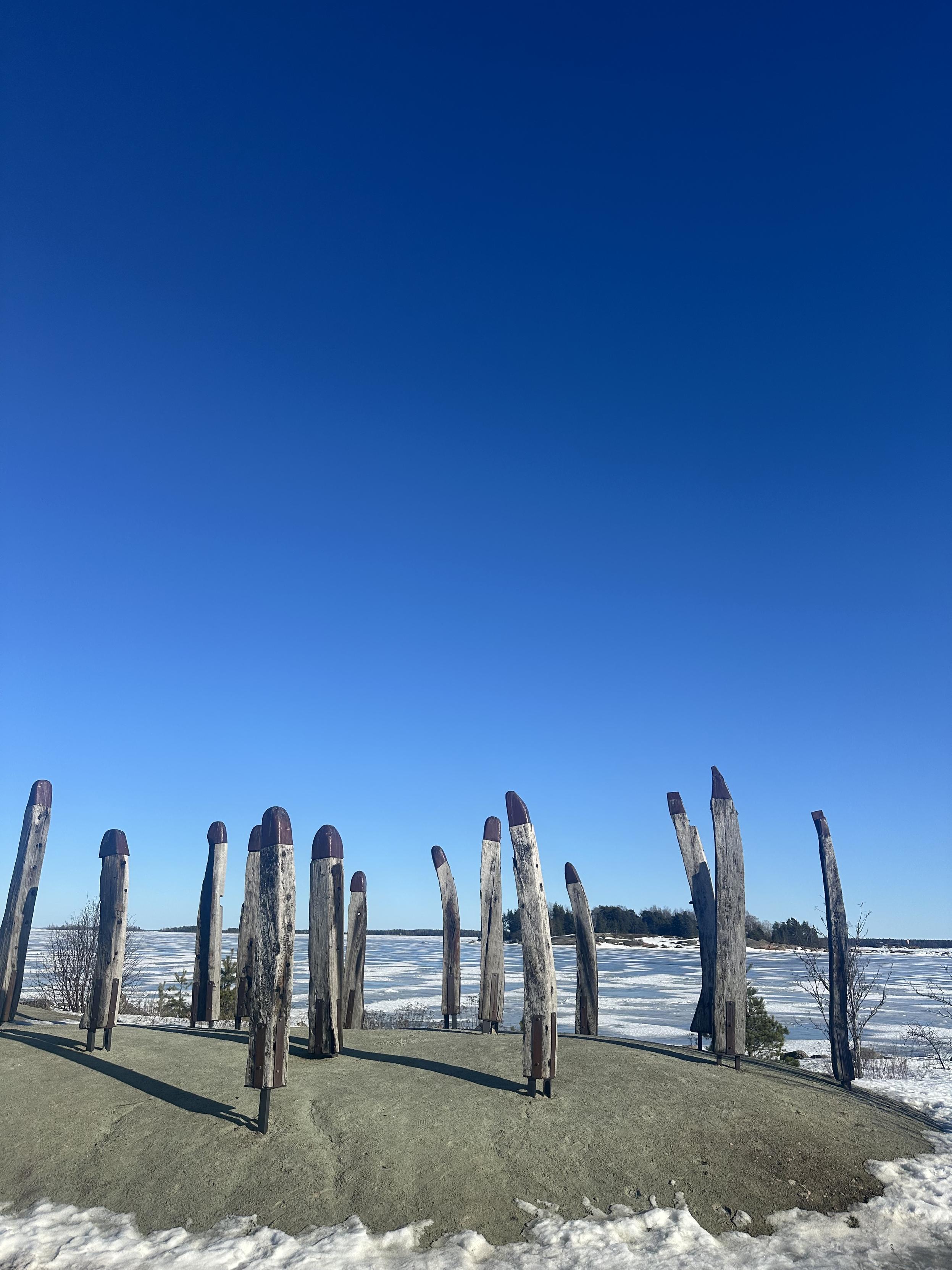 Puisia pylväitä metallisilla kärjillä kurottaa kohti sinistä taivasta. Taustalla jäinen meri ja siitä nouseva saari