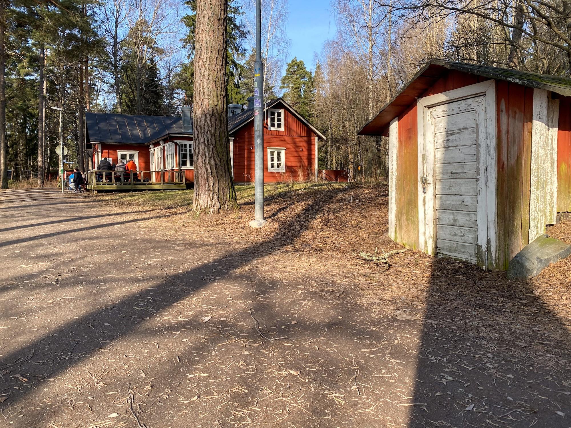 Hiekkaisen kävelytien oikealla puolella lähinnä maakellarin tai varaston sisäänkäynti. Vähän matkan päässä järeän männynrungon takana punainen kahvilatupa. Sen terassilla istuu ihmisiä. Aurinko luo jyrkkiä puunrunkojen varjoja tien poikki.