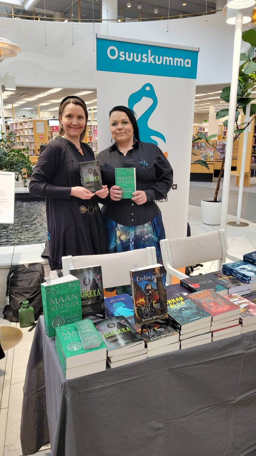 Kuvassa kirjailijat Nyström ja Morre, jotka pitelevät kirjojaan Osuuskumman myyntipöydän takana. Heidän takanaan Osuuskumman mainos-roll-up ja Pasilan kirjaston suihkulähde.