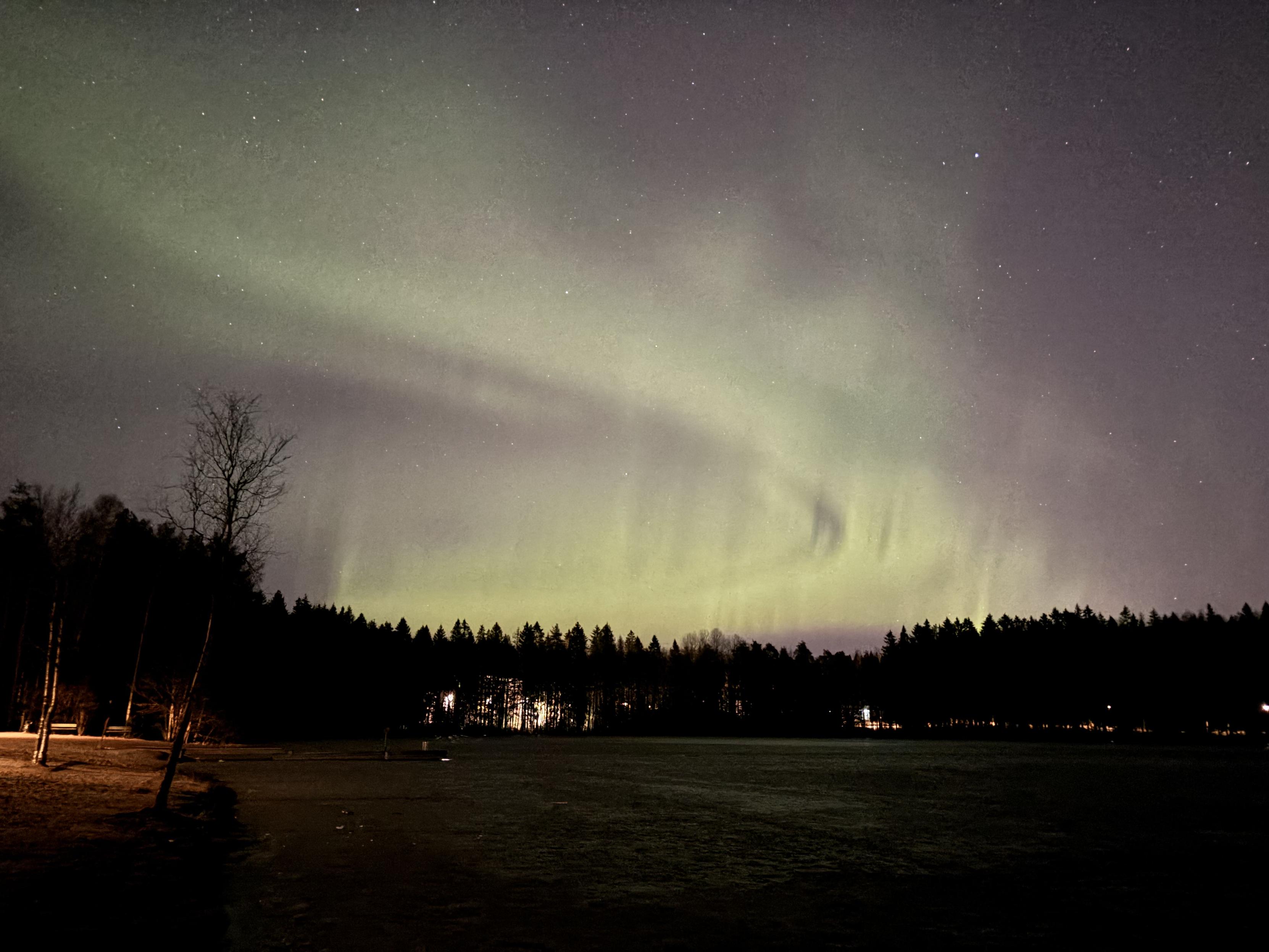 Härskisti ylivalottunut kuva revontulista  pienen järven yllä. Reposet ja järvenjään tekstuuri erottuvat kivasti, mutta taivas on hailakan punaharmaa.