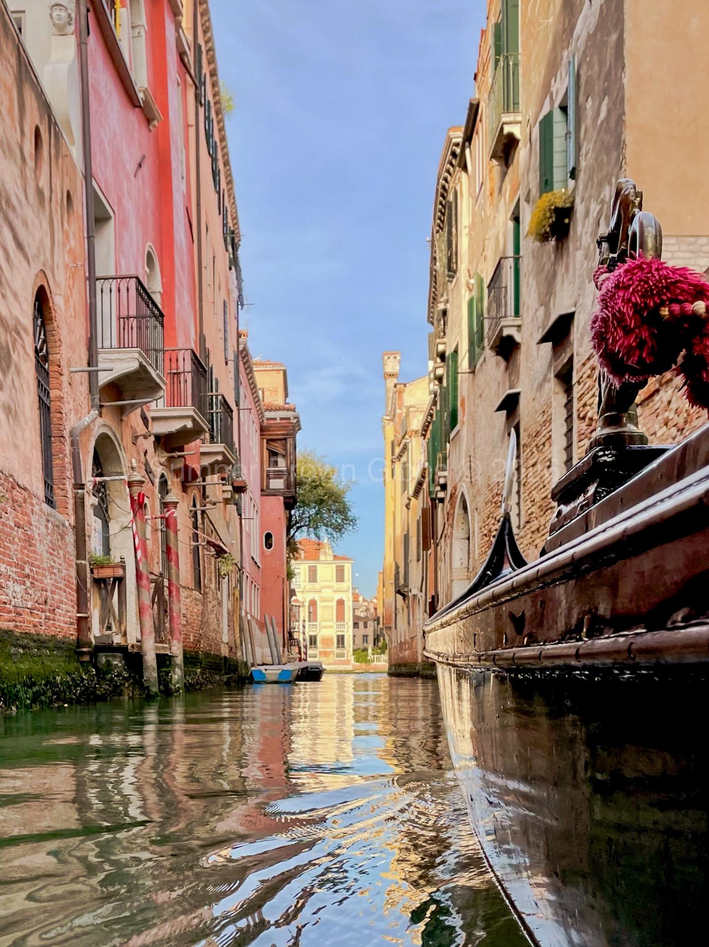 view from a gondola in colorful venice canal with camera hovering over water; side view of gondoal is visible