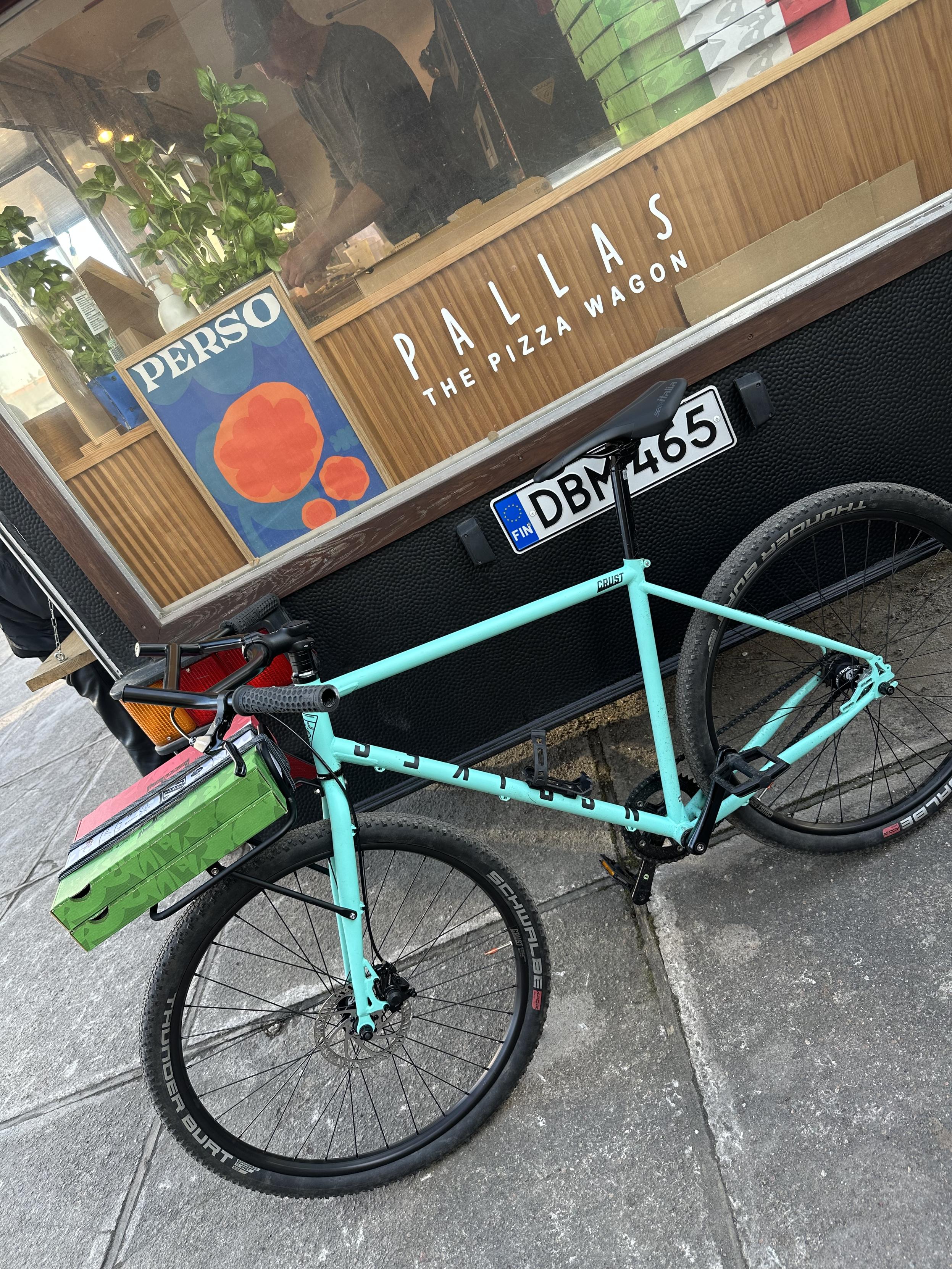 Fiksipyörä etutarakalla nojailee Pallaksen pizza wagonia vasten. Tarakalla on kaksi pizzalaatikkoa.