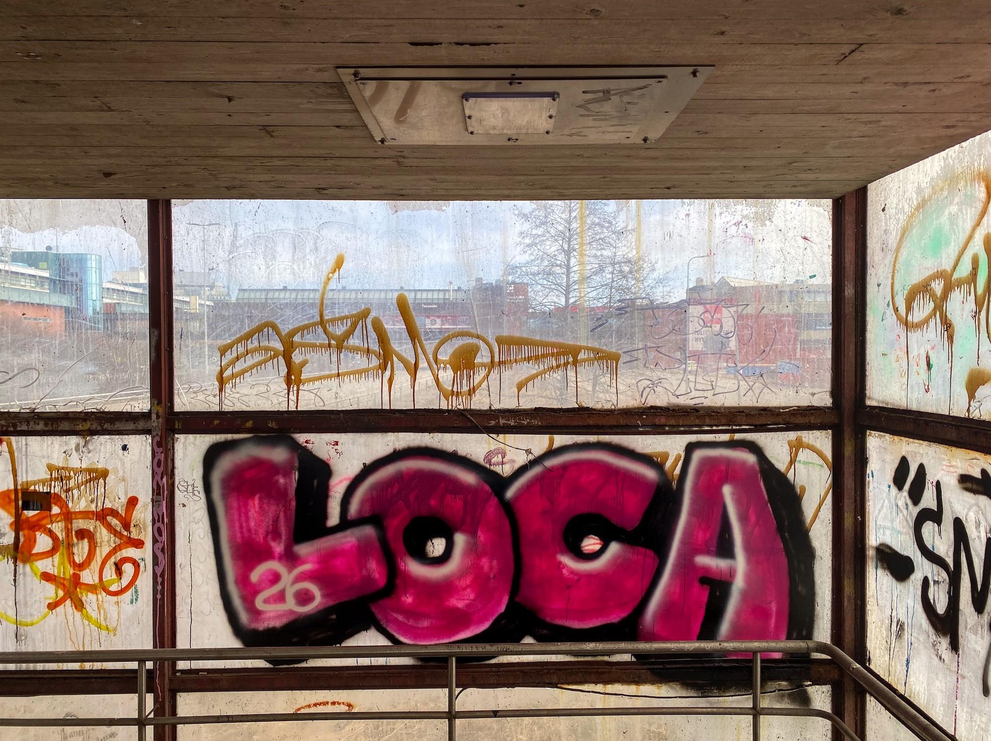 Näkymä rähjääntyneestä portaikkotornista graffitin peittämien ja sameiden pleksilasien läpi kohti monikaistaisen tien ylle rakennettua ostoskeskuksen osaa. Keskimmäisen ikkunaruudun täyttää suuri teksti "LOCA".