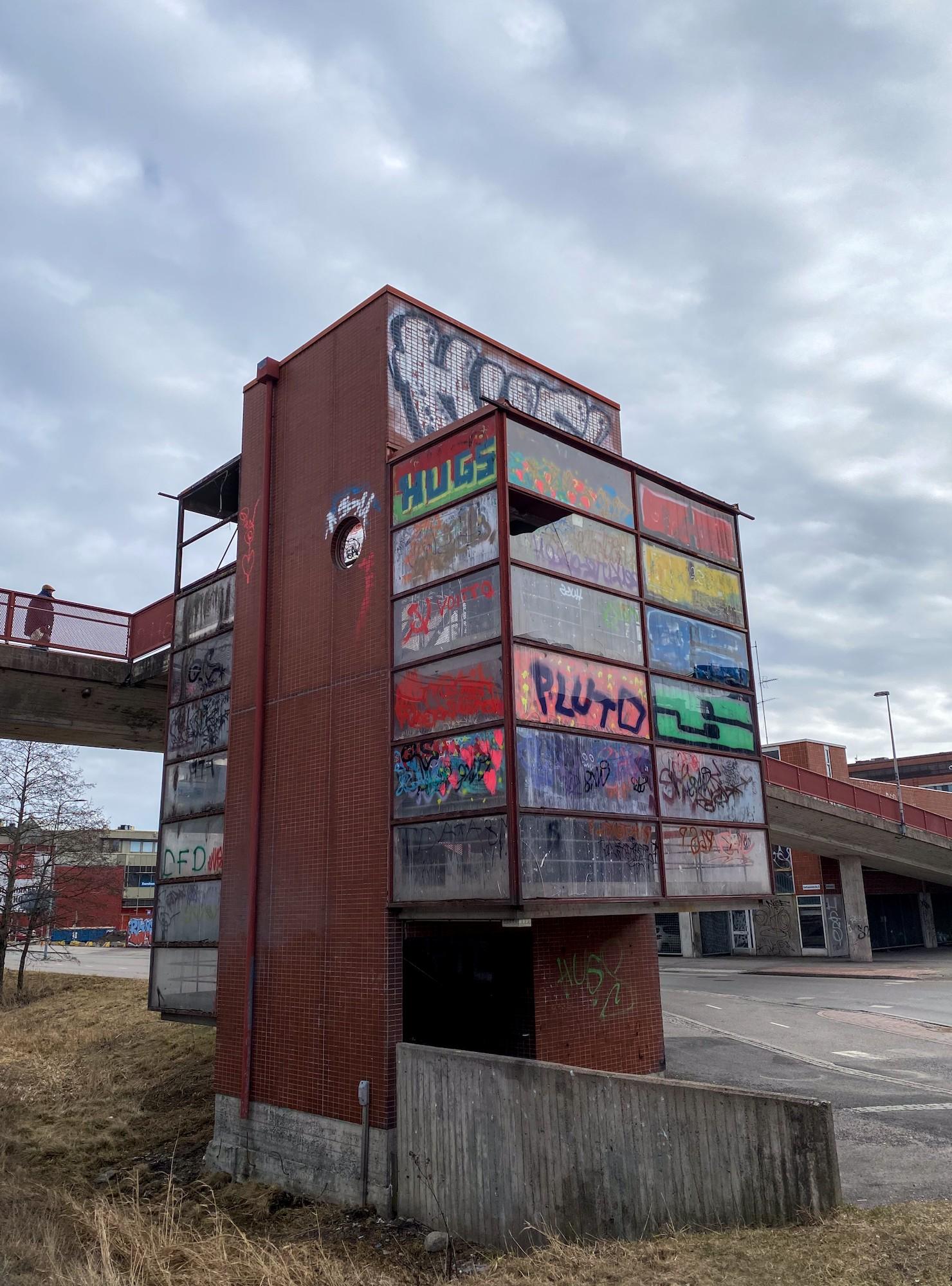 Kävelysillalle alapuoliselta tieltä johtava portaikkotorni kuvattuna alaviistosta kohti pilvistä taivasta. Tornin pleksilasiset seinät ovat monenväristen graffitien peitossa.
