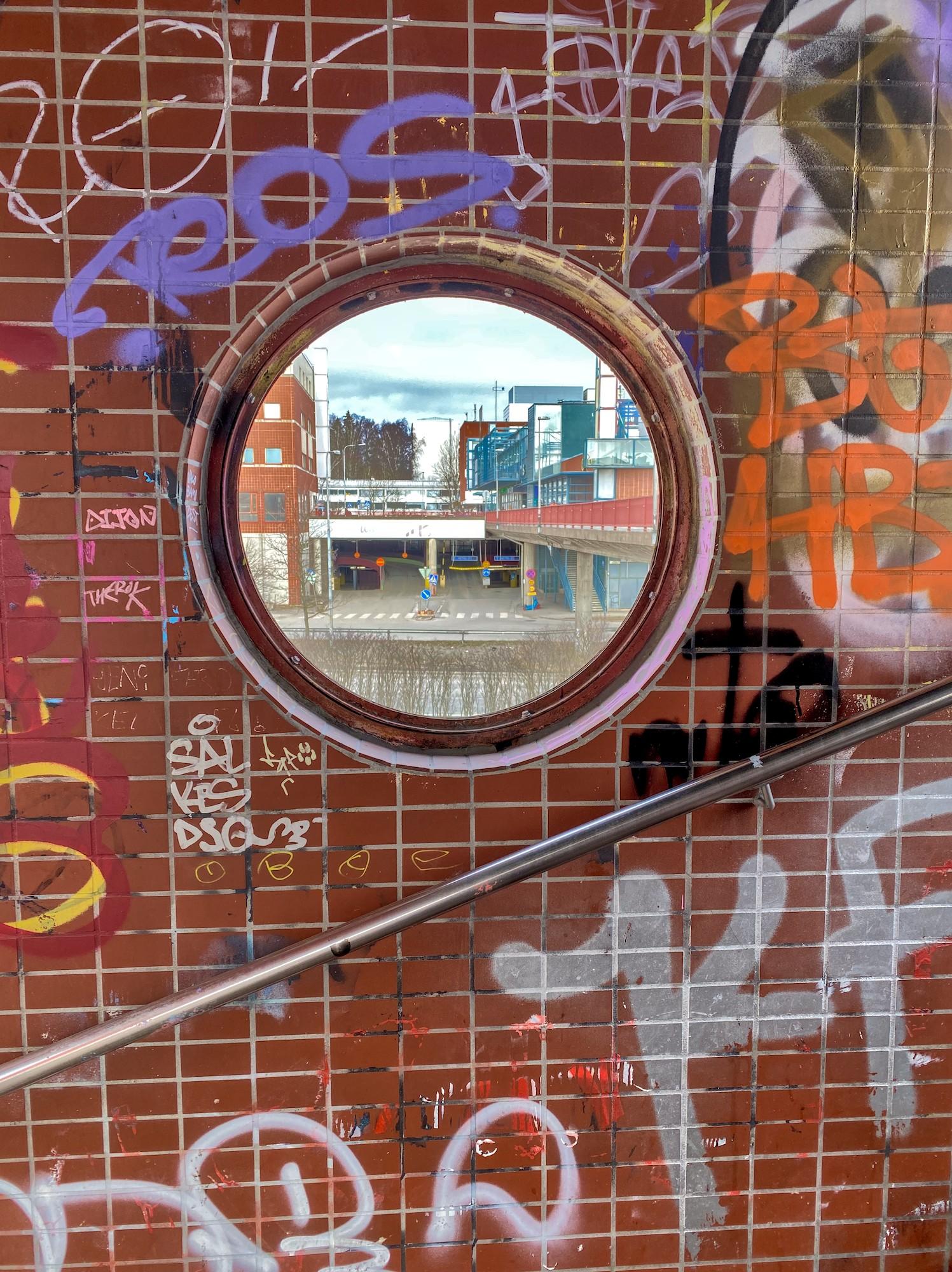 Portaikon punatiiltä jäljittelevä laattaseinä, joka on graffitien peitossa. Keskellä on pyöröikkuna, josta näkyy etäämmällä tien takana ostoskeskuksen rakennuksia ja kerrostalojen päätyjä.