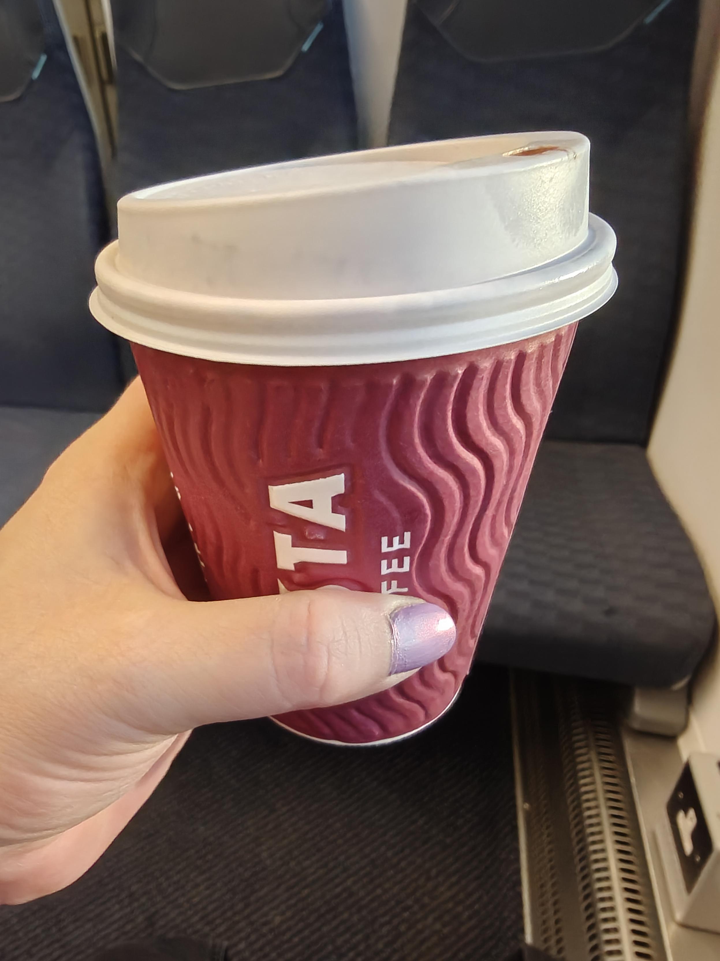 My hand holding out a fresh morning mocha in a red Costa Coffee cup on public transport. I'm showing off the colour of my iridescent purple polish on my thumb nail.