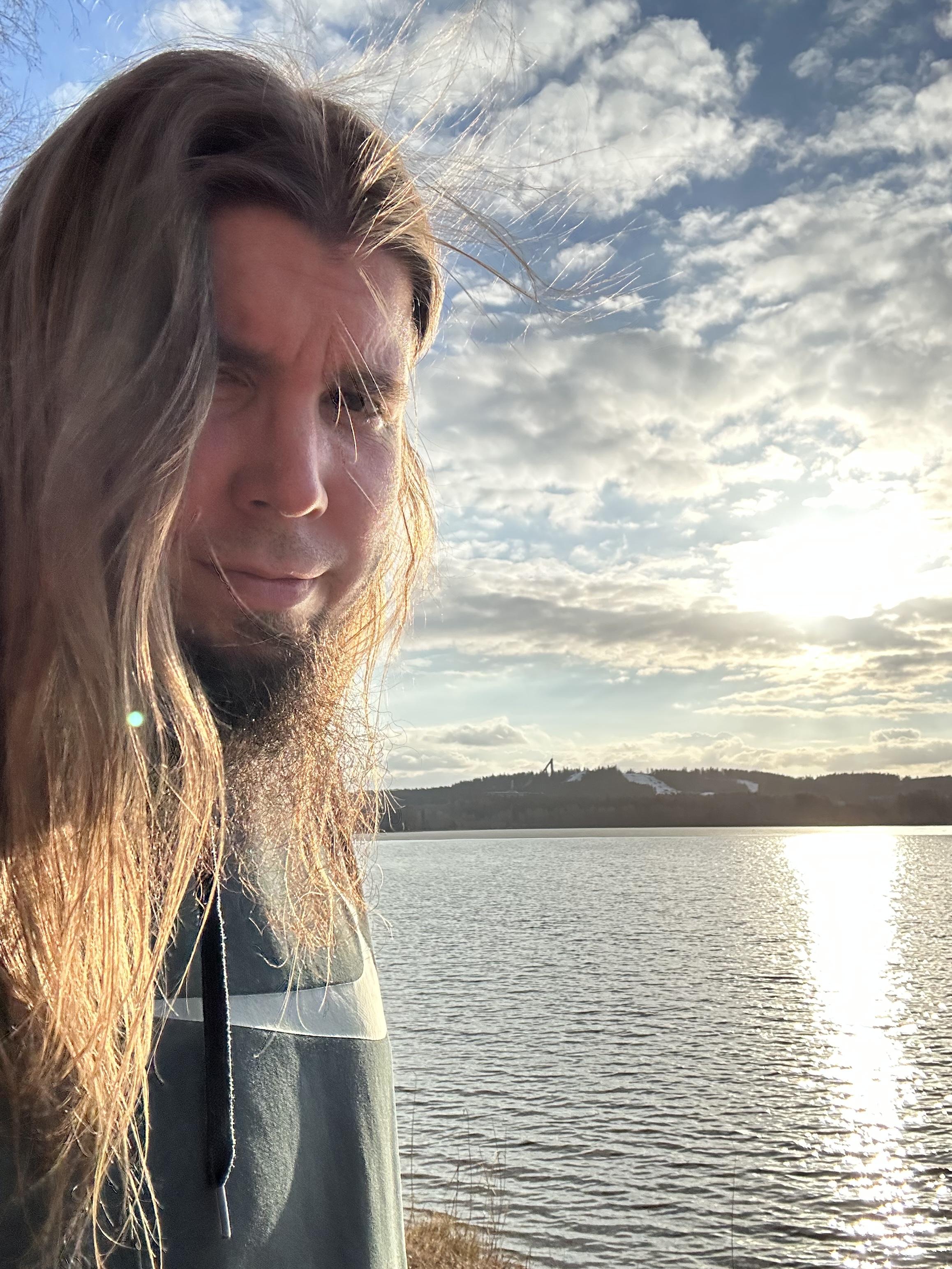 Mies järven rannalla, sää aurinkoinen ja taivas pilvinen, taustalla järvi ja kaukana mäkiä.