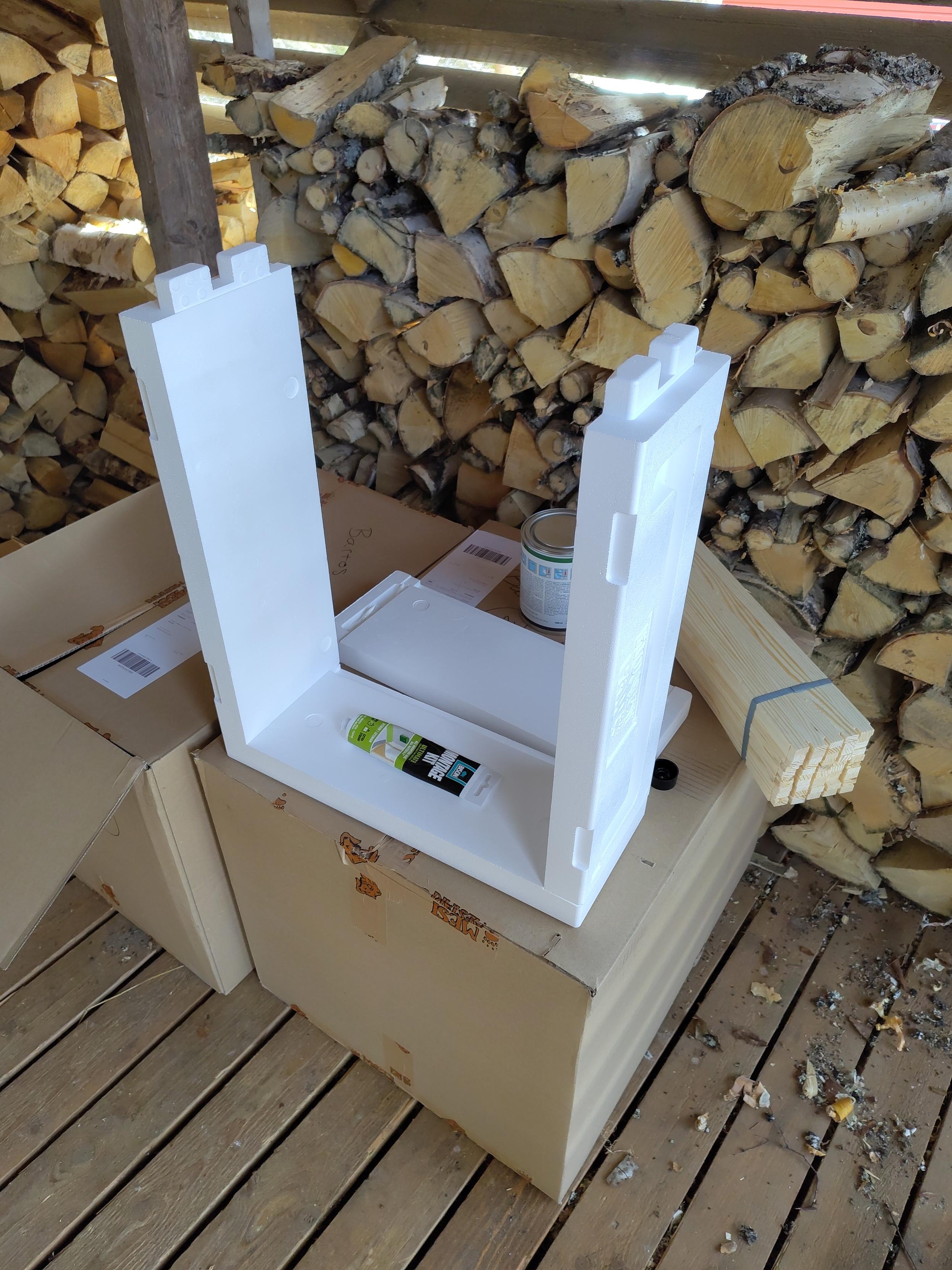 Mehiläispesälaatikko kasausvaiheessa pahvilaatikon päällä. Pahvilaatikossa on lisää pesälaatikoita. Taustalla puupinoja.