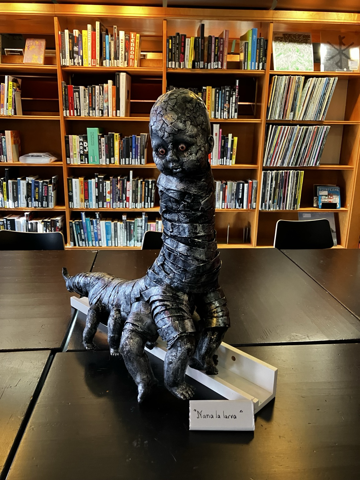 Vauvanpäistä, monijalkaista, harmaanhohtoista luikeroa esittävä veistos kirjahyllyjen edessä.