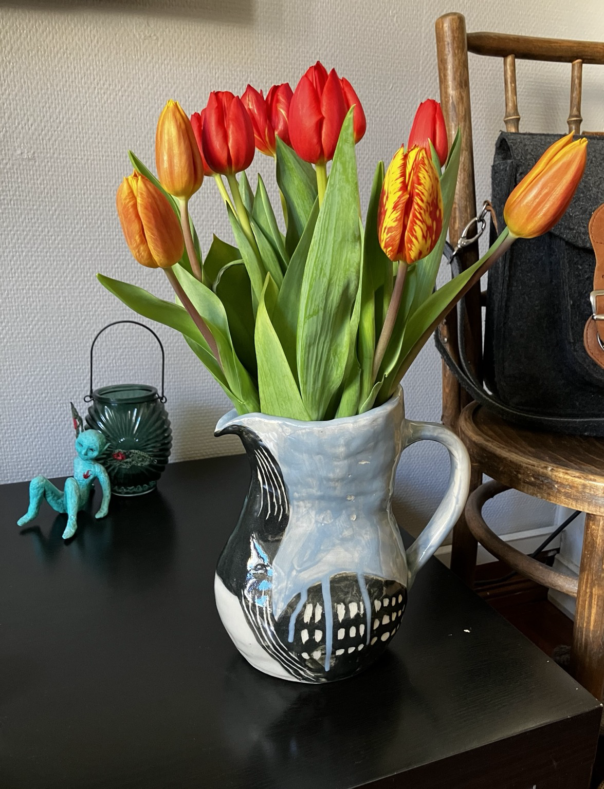 Punaisen ja keltaisen kirjavia tulppaaneja keraamisesta kannussa, jonka väritys muistuttaa kuikan ominaisväritystä. Taka-alalla vihreä kynttilälyhty, johon nojaa turkoosi keijufiguuri.