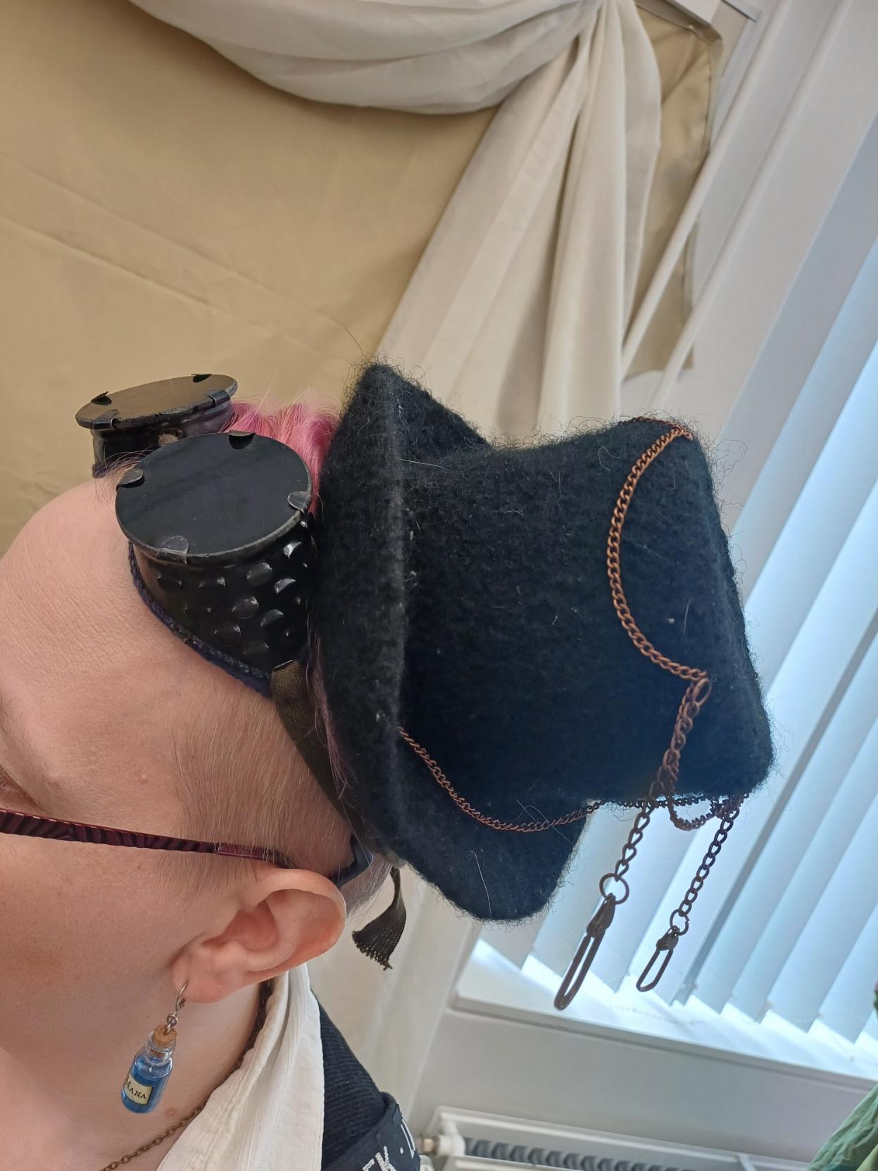 Lähikuva mustasta silinterihatukkeesta lyhyttukkaisen naisen päässä. Naisella lisäksi tummat suojakakkulat otsalla, silmälasit sekä mana-pullo korvakoruna.