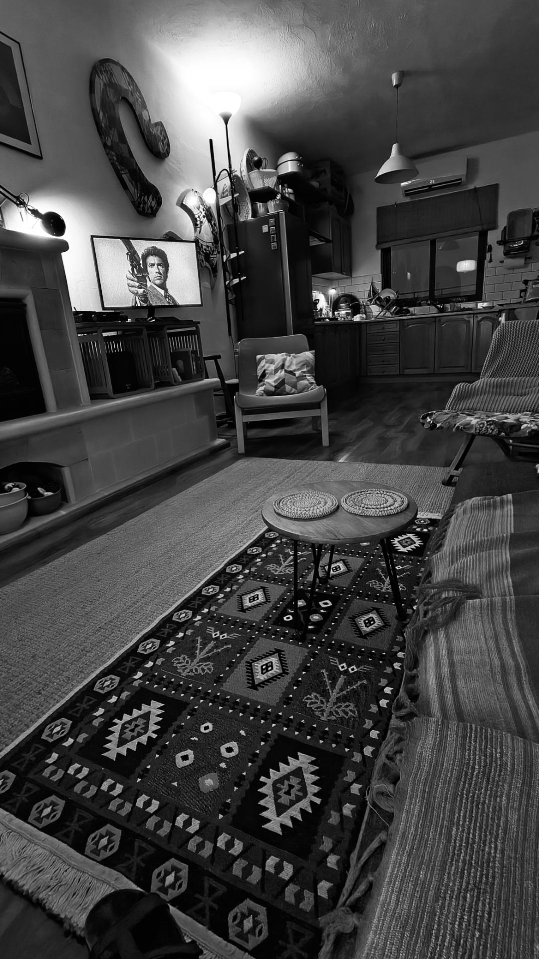 Evening view of the living room, Magnum Force on the media screen. Art and wall lights, patterned rug, stone fireplace, low lighting, a kitchen area beyond.