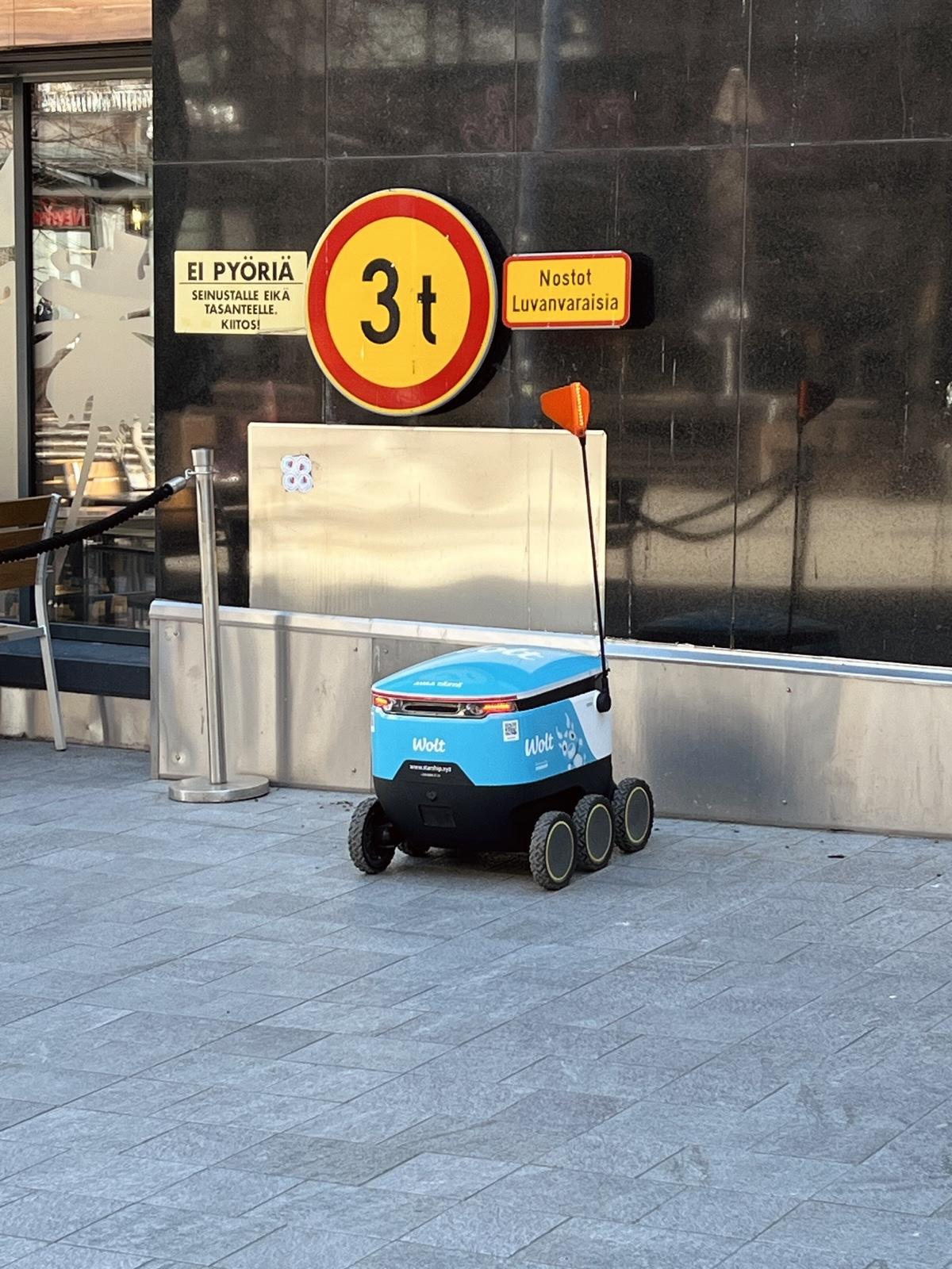 Kuljetusrobotti
Woltin sinivalkoinen kuljetusrobotti seisoo kivipäällysteisellä jalkakäytävällä rakennuksen seinustalla. Robotin kyljessä näkyy selvästi Wolt-logo. Robotissa on kuusi pyörää ja oranssi näkyvyystikku. Taustalla liikennemerkki ja kyltti "Ei pyöriä seinustalle".