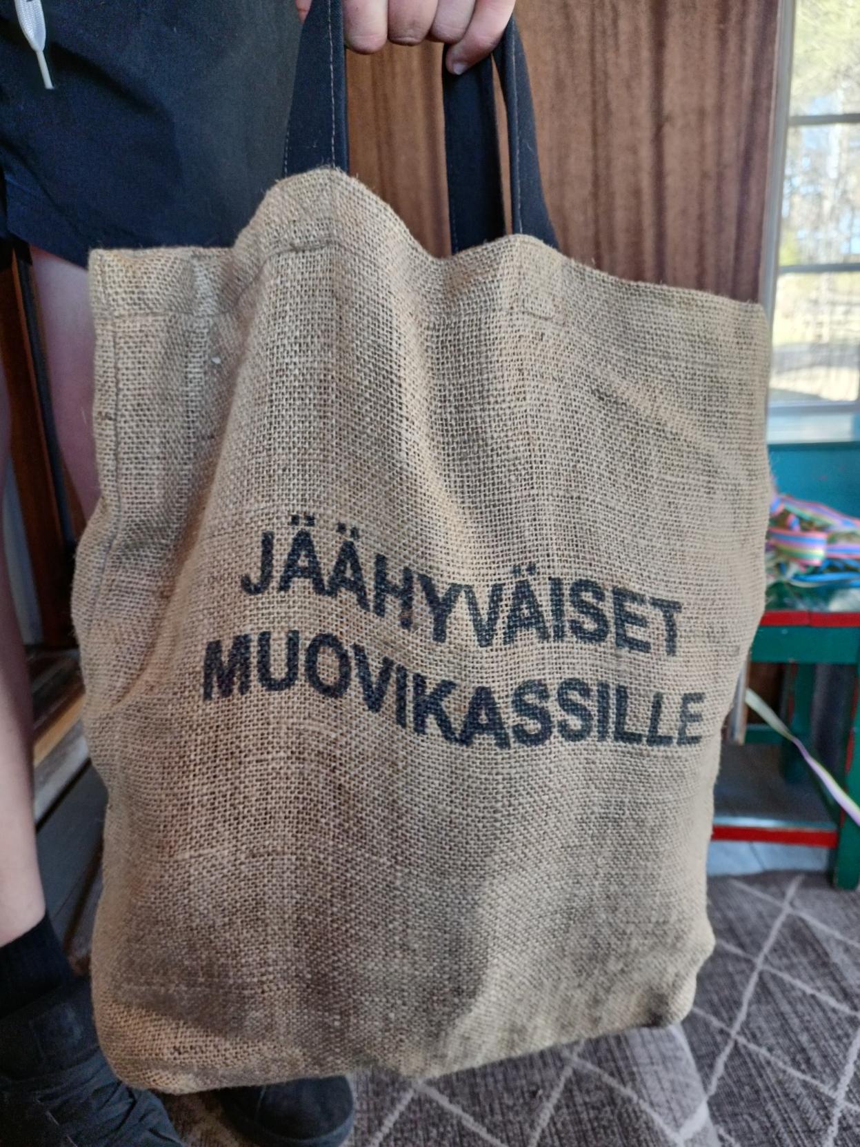 Kuvassa henkilön kädessä roikkuu juuttikankainen kassi, johon mustalla painettu teksti "jäähyväiset muovikassille".
