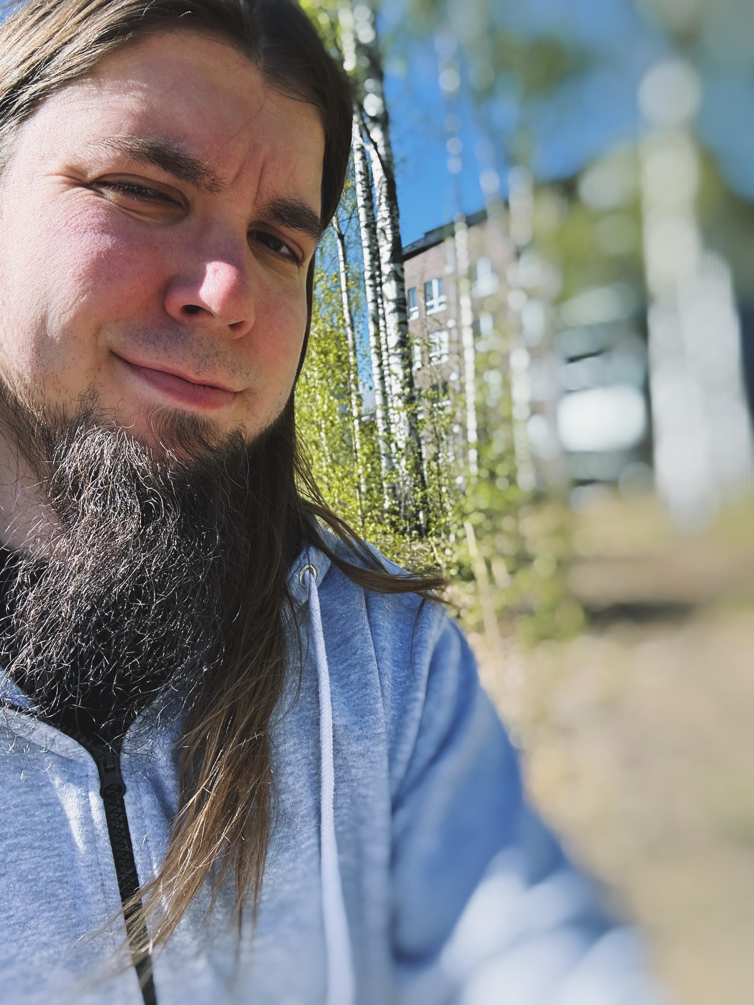 Rolle pihalla, pitkät tummat hiukset ja parrakas, hän hymyilee ja taustalla on luontoa ja rakennuksia, päivällä kirkkaassa auringonvalossa.