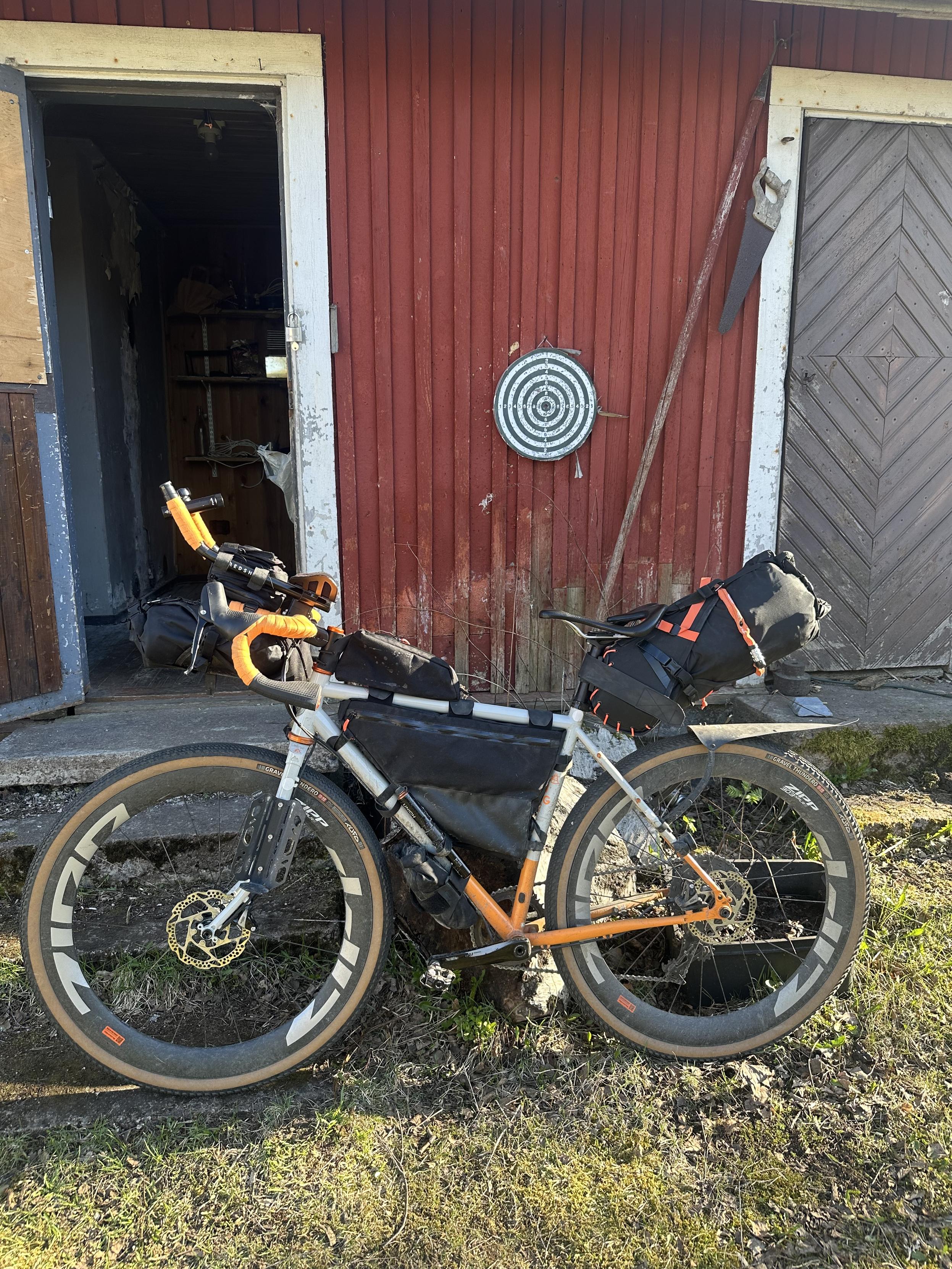 Harmaaoranssi retkipyörä täynnä laukkuja nojaa koivupölkkyyn punaisen piharakennuksen edessä.