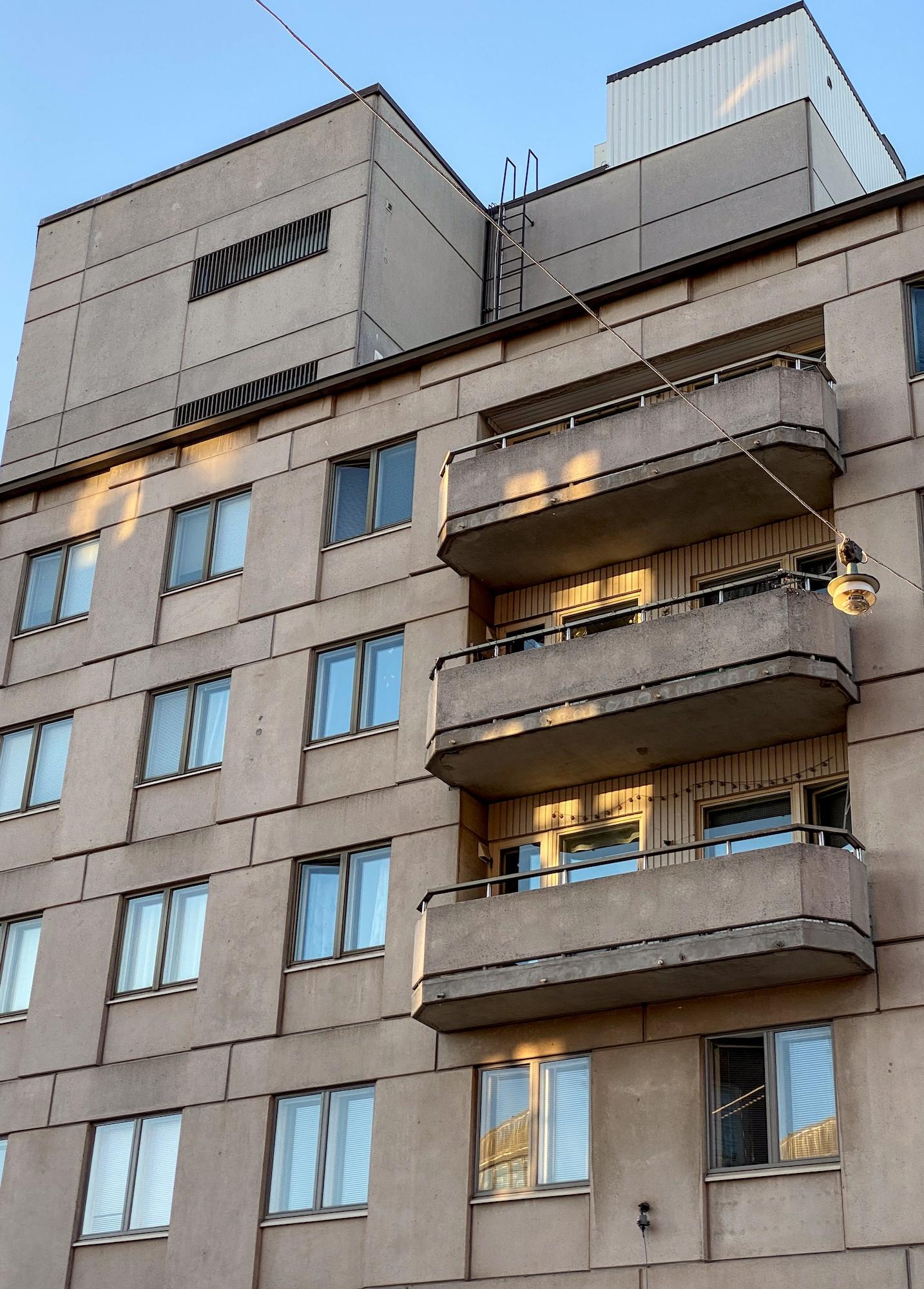 Harmaa betonikerrostalo alaviistosta. Vasemmalla neljässä kerroksessa samanlaisia kaksiruutuisia ikkunoita. Oikealla kolmessa kerroksessa ulkonevia parvekkeita, joiden päädyt ovat hieman viistot. Asuinkerrosten yläpuolella katolla suuria kulmikkaita tiloja.