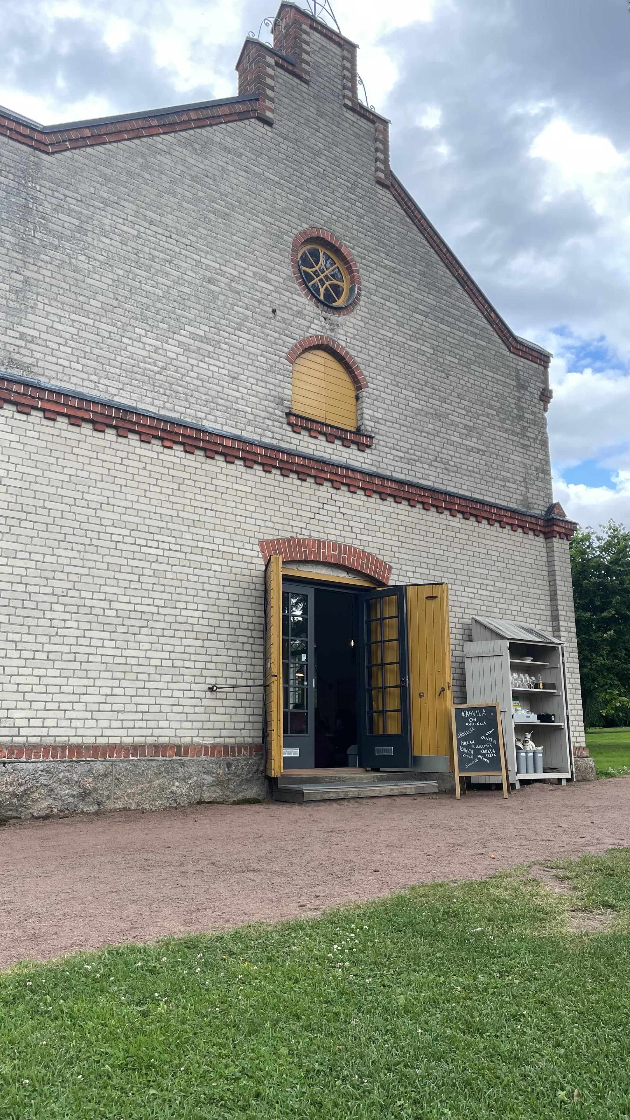 Myllymakasiinin pääty, harmaatiilinen rakennus, jossa punatiiliset koristeet ja ovet avoinna, sillä sisällä on kahvila. Oven vieressä on liitutauluständi ja astiapalautukseen käytetty hylly.