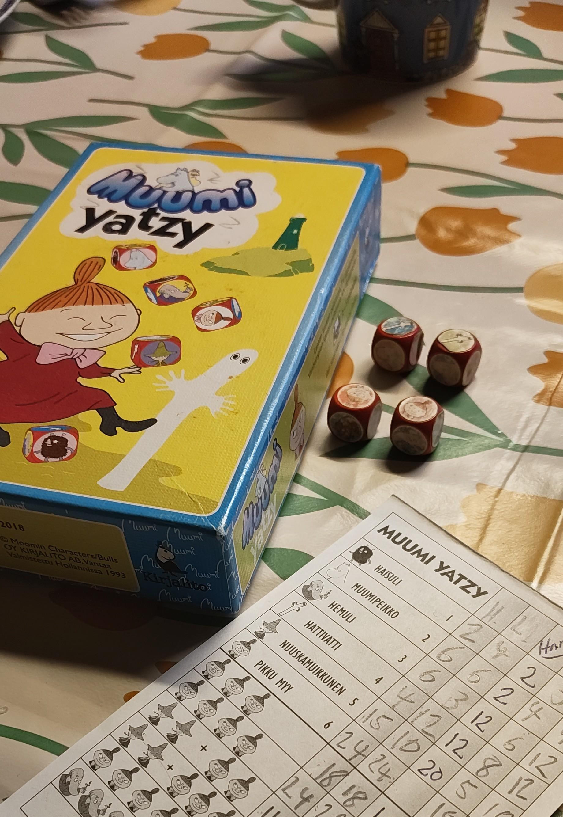 Kukkakuvioisella pöytäliinalla Muumi Yatzy pelin pistelomake, neljä noppaa, joissa kuluneet muumihahmotarrat, sekä pelin laatikko, jonka kannessa iloinen Pikku Myy, hattivatti, majakka ja lentävät muumikuvioiset nopat.