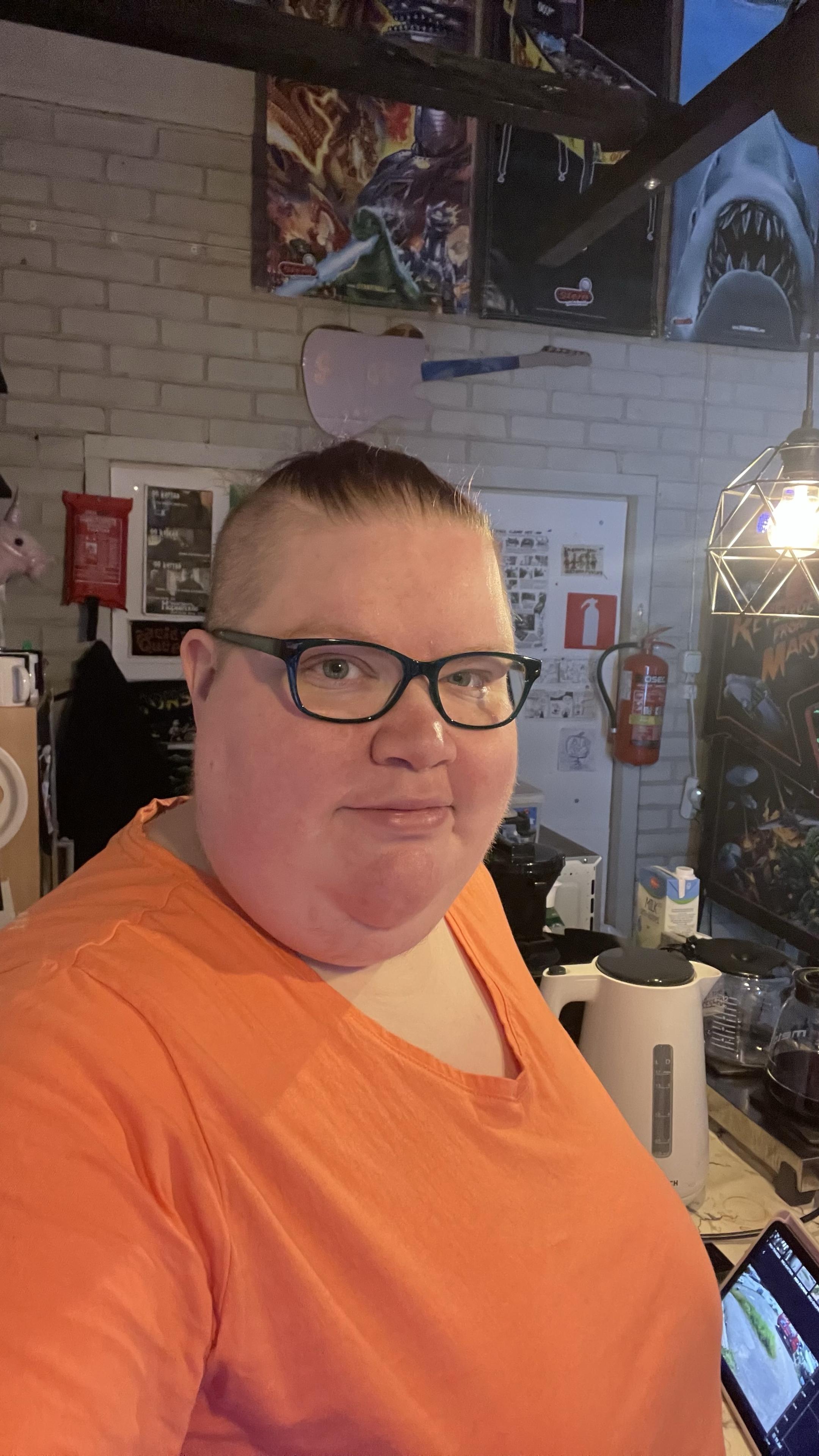 A photo of me in my cashier spot at our arcade. There are all kinds of things in the background, such as pinball themed banners up on the wall, some of my cross stitchings on a door and the left side of Revenge from Mars pinball machine on the right side of the photo