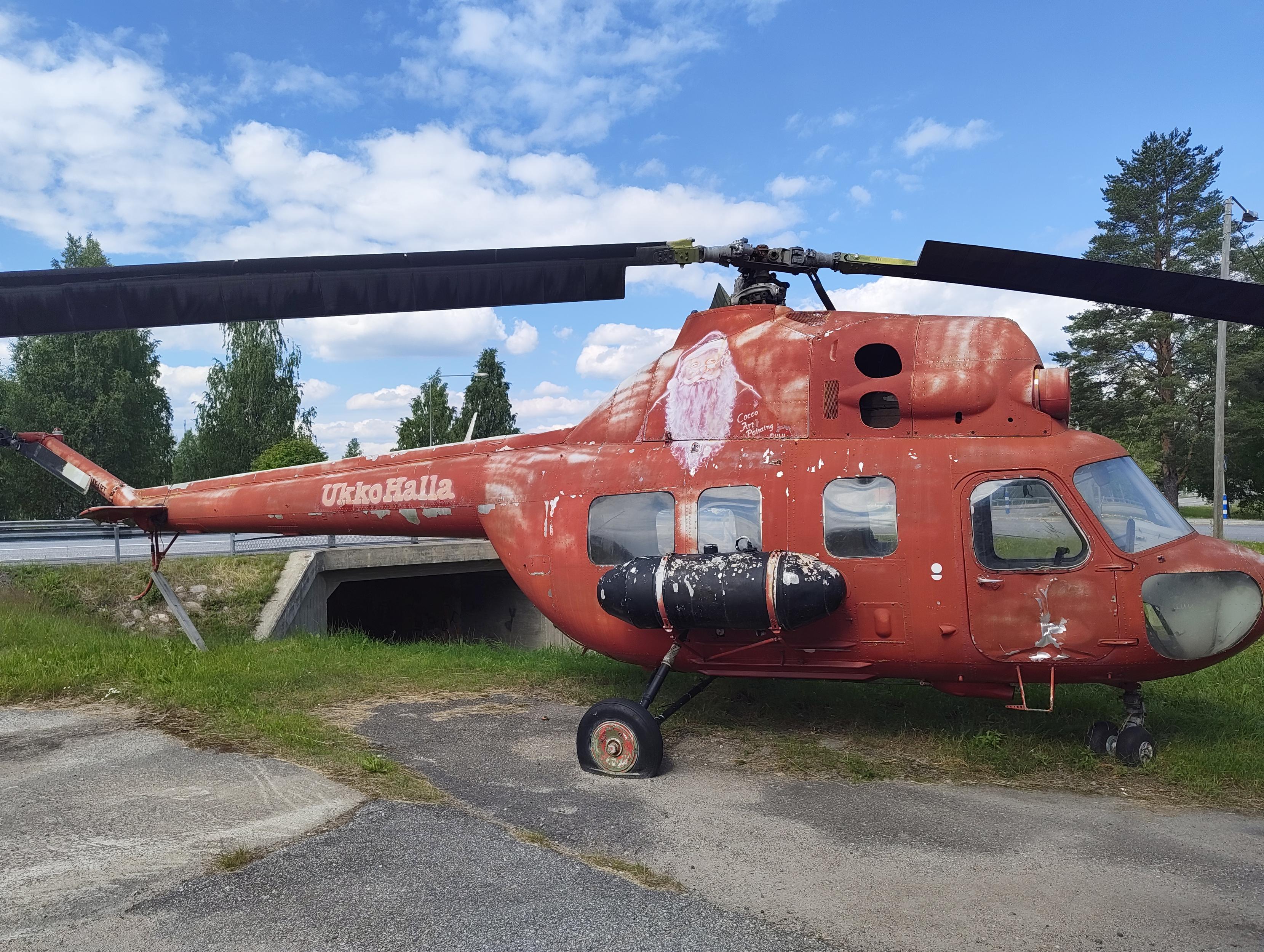 Punainen helikopteri vanhan huoltoaseman pihalla. Kopterissa joulupukin kuva ja teksti UkkoHalla, joka on pieni laskettelukeskus Hyrynsalmella.