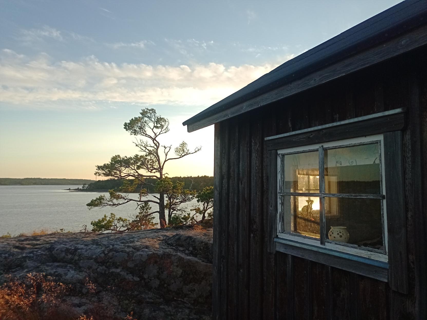 Ilta-aurinko paistaa pienen, tervatun mökin neliruutuisten ikkunoiden kautta mökin läpi.
Mökki seisoo kalliolla ja sen vieressä kasvaa pieni mänty. Taustalla näkyy Itämeri ja Boxönudde.