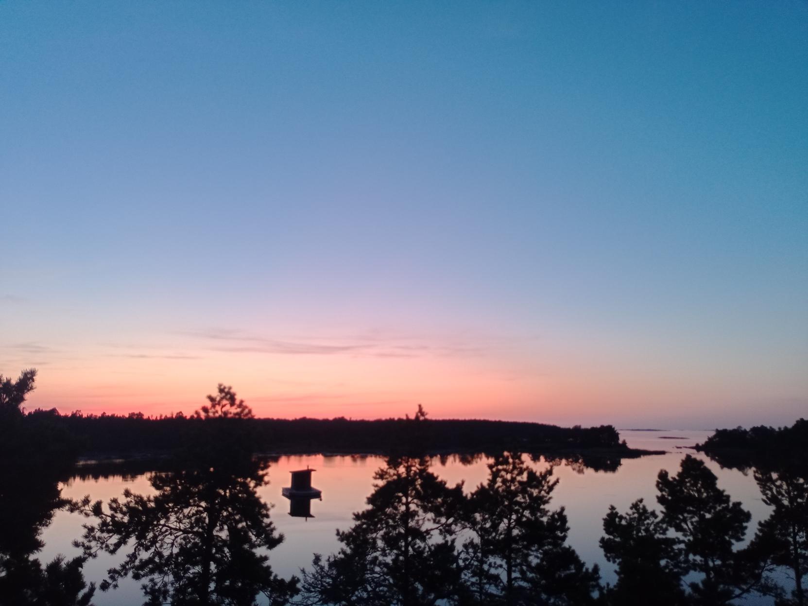 Aurinko on laskenut ja taivas liukuu alhaalta ylöspäin punaisesta oranssin kautta siniseen.
Merenlahti on täysin tyyni ja taivas, puut ja lahdella kelluva saunalautta peilautivat vedestä.