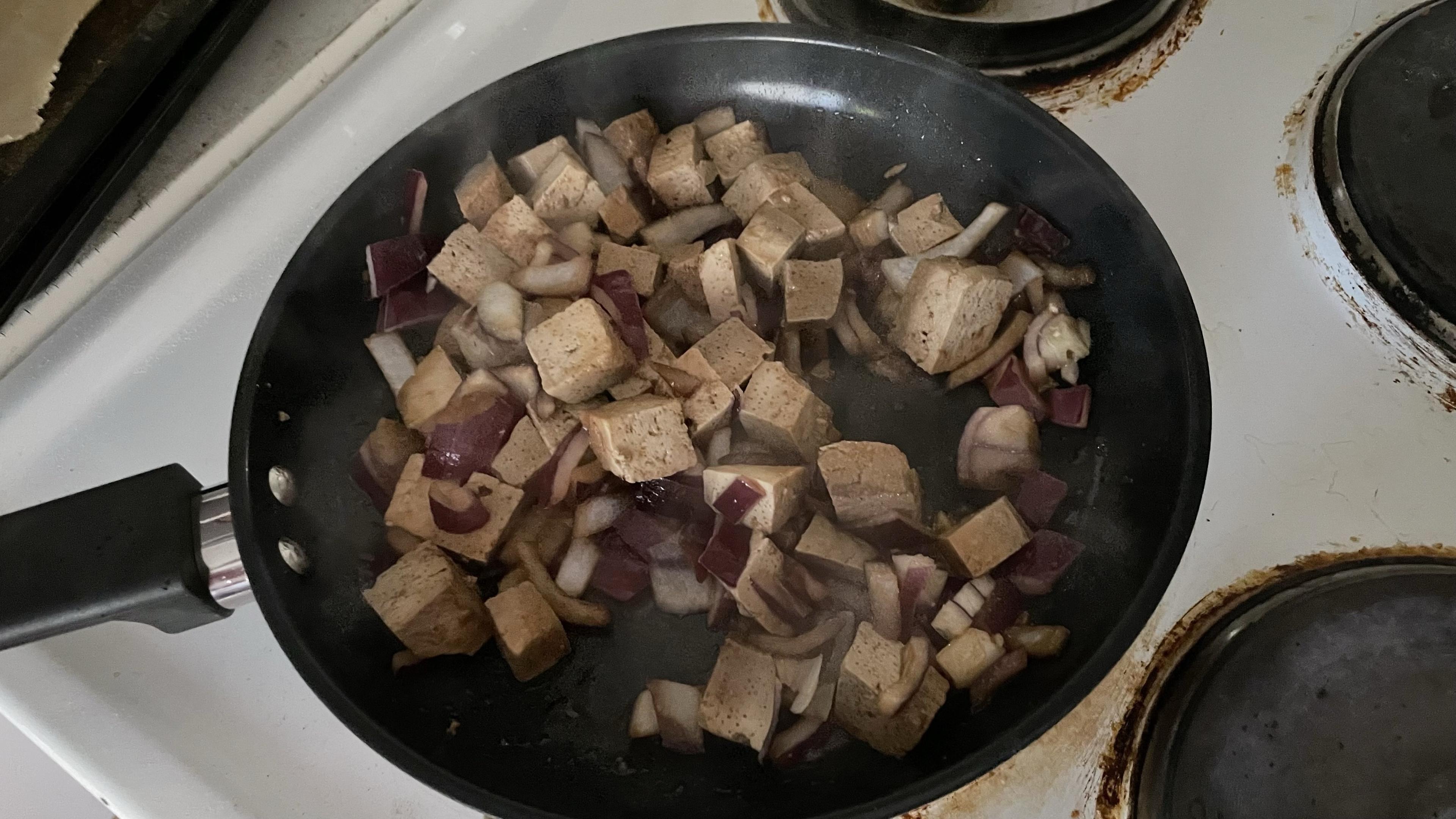 A frying pan with cubed tofu and chopped onions frying in it.