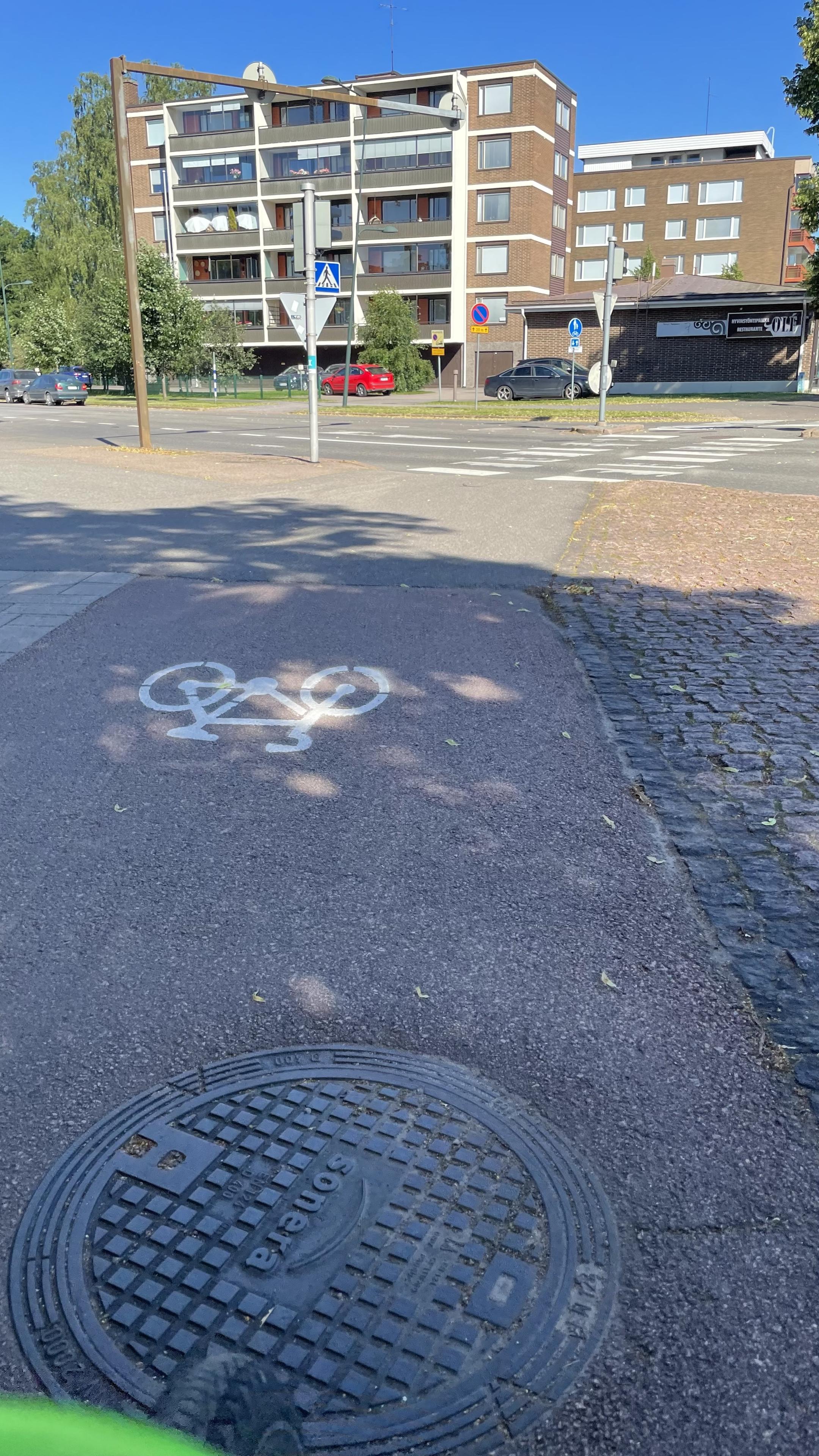 A shot that I took with my phone attached to my bike's handle bar. I am cycling on the bike lane, coming up to a cross roads with lights. On the other side of the road a couple of apartment buildings and some parked cars are visible.