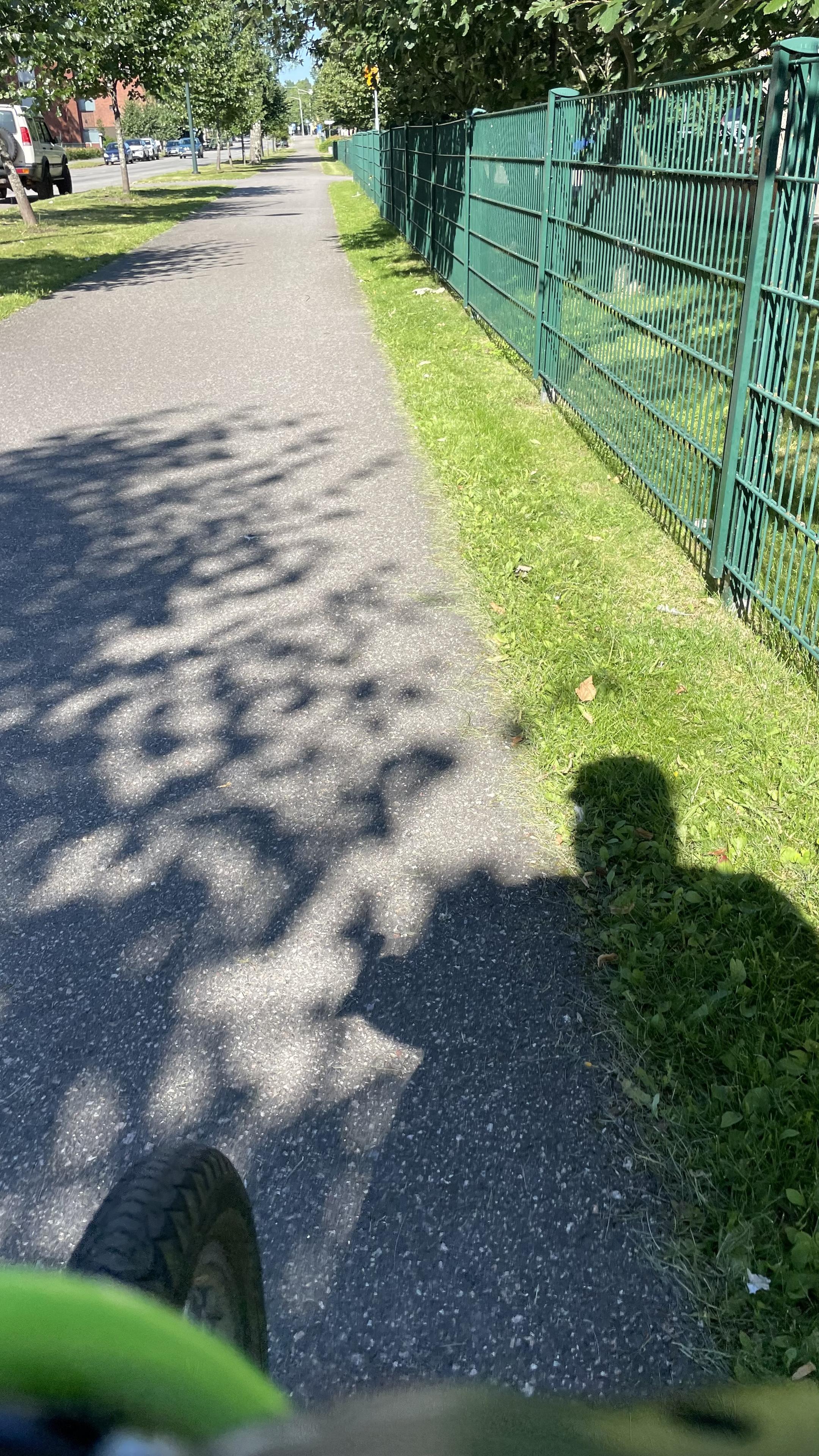 I am riding past the apartment buildings, which would be on the right, behind the green fence visible in the picture. After the fence a public park is coming up. On the ground my shadow and the shadow of a tree on my left are also visible.