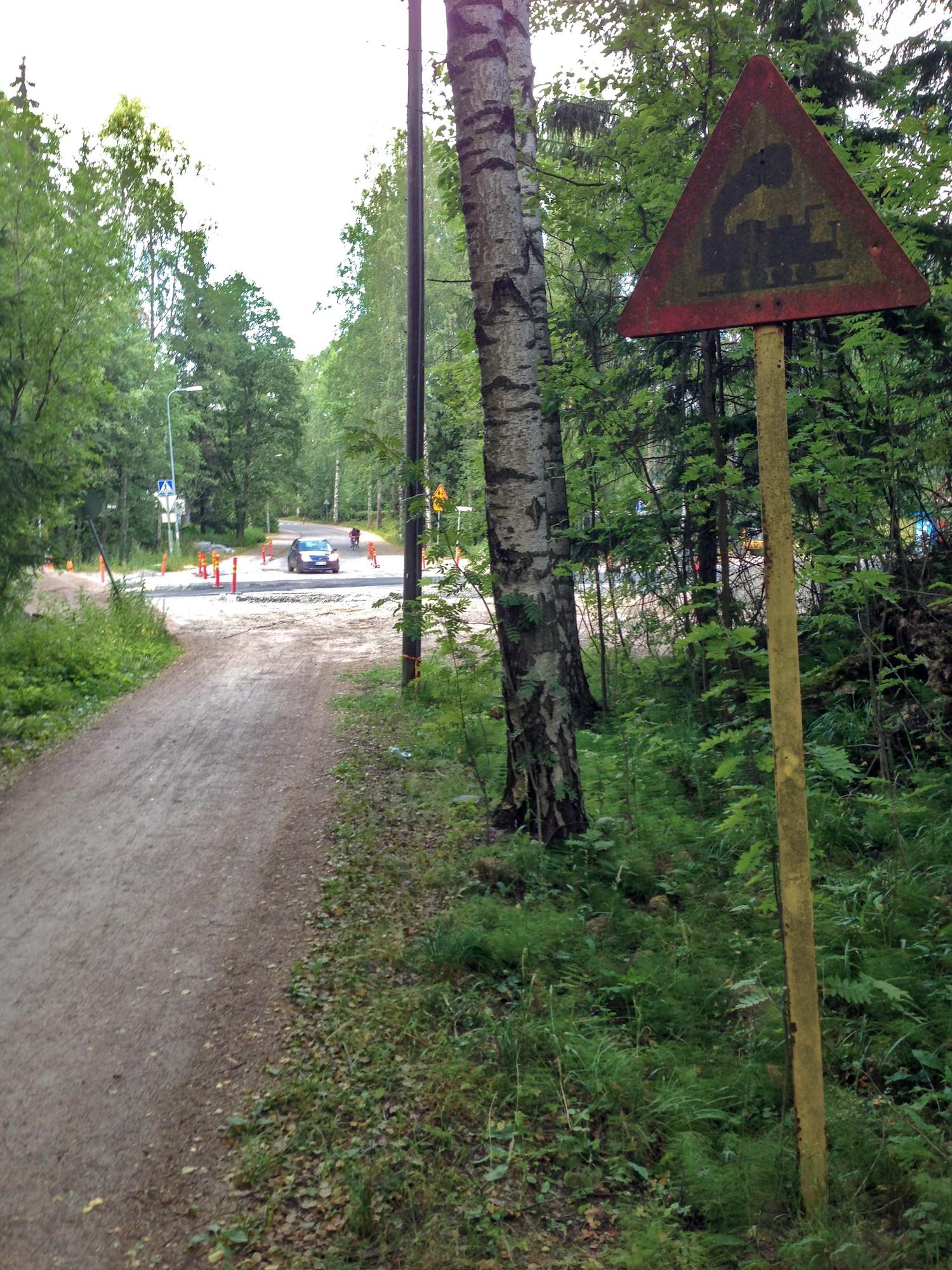Sorapintainen kapea pikkutie johtaa lehtipuiden välistä isomman tien risteykseen. Risteävällä isolla tiellä on työmaan ohjausmerkkejä. Suoraan risteyksen toisella puolella jatkuvalta tieltä kääntyy auto täältä katsottuna oikealle. Lähinnä pikkutien oikealla puolella on rautatien tasoristeyksestä varoittava liikennemerkki.