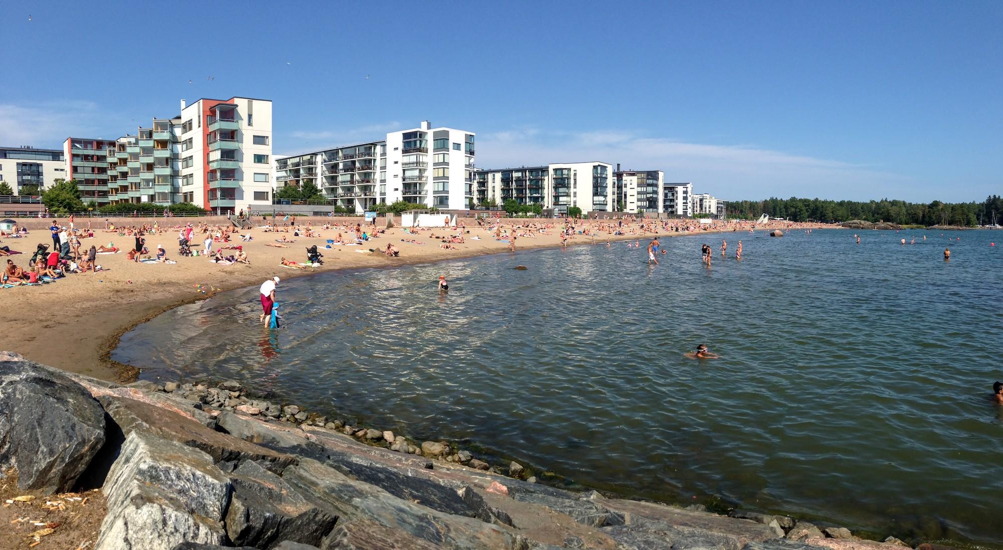 Iso hiekkaranta täynnä ihmisiä. Etualalla kivettyä rantaa, joka vaihtuu kuvan vasemmassa reunassa hiekkarannaksi ja kaartuu etäisyyteen kohti kuvan oikeata yläosaa. Vettä on puolet kuva-alasta. Ihmisiä on vedessä ja rannalla, he istuvat, makaavat, uivat ja kahlaavat. Hiekkarannan takana on rivi valkoisia 4–5-kerroksisia asuintaloja.