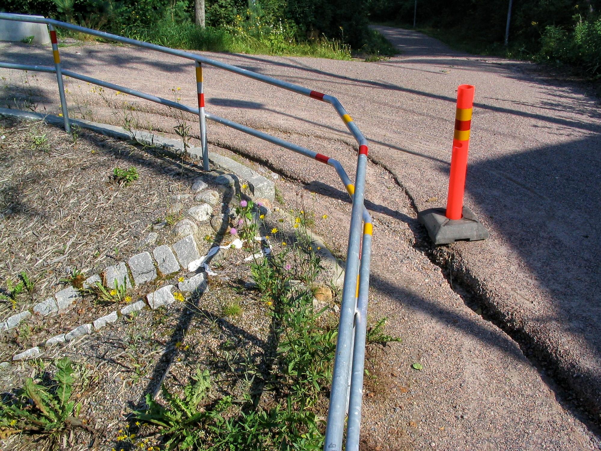 Sorapintaisen kävelytien risteys. Kuvaajan takaa oikealta nouseva tie haarautuu vasemmalle ja suoraan eteenpäin. Vasen haara jatkaa ylämäkeen ja häviää kuvan vasempaan yläkulmaan. Tietä reunustaa vasemmalla metallinen kaide, ja sen luiska viettää selvästi alas vasemmalle. Risteyksestä lähtee tiestä pois päin kivistä ladottu kouru, jonka alku on tienpintaa korkeammalla. Vasemmalta tietä pitkin valunut vesi on uurtanut soratiehen syvän jäljen, joka jatkuu tietä pitkin ulos oikeasta alakulmasta. Uurteen reunaan on pystytetty muovinen varoitustolppa.