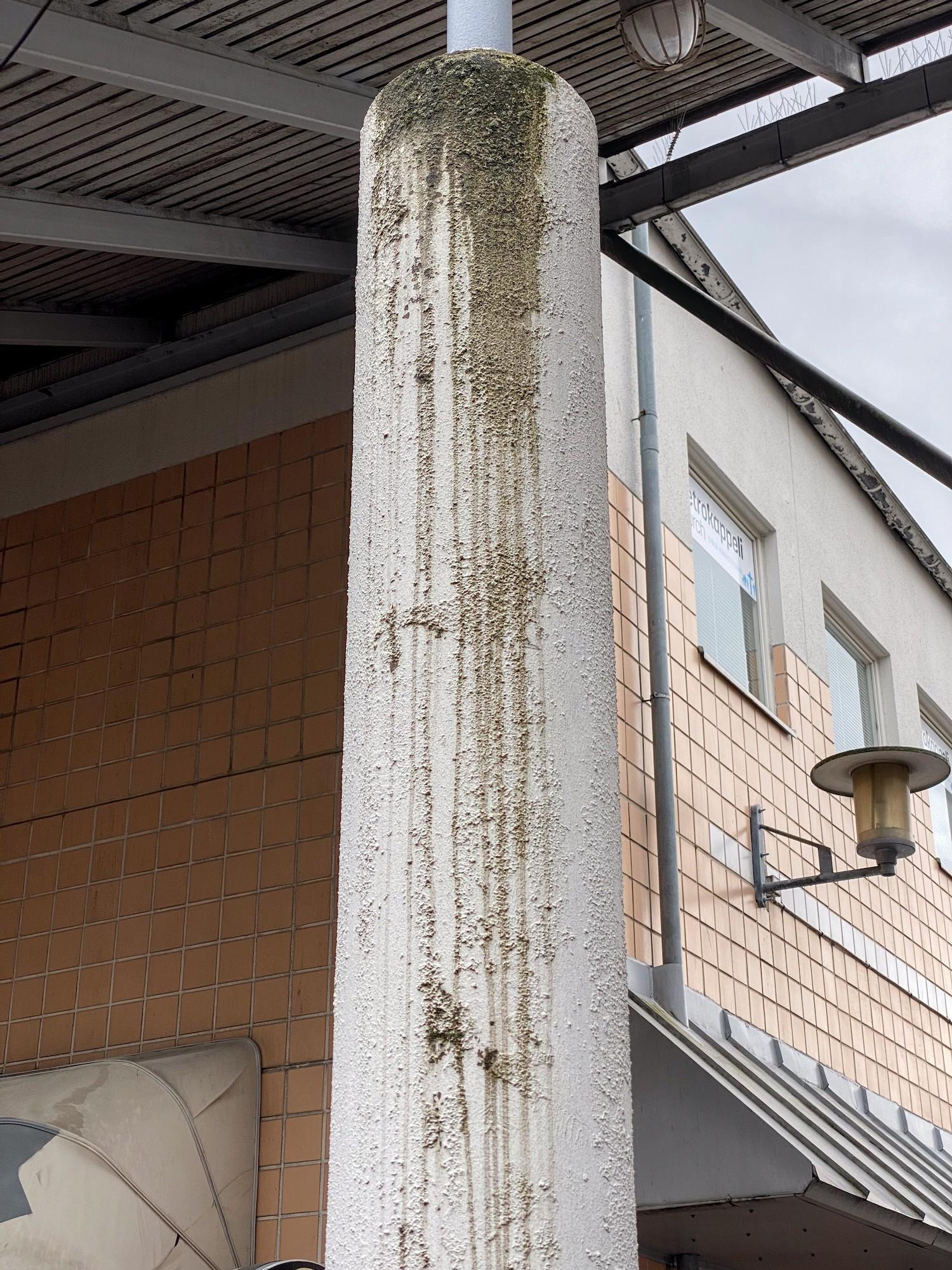 Valkoiseksi rapattu pylväs, jonka päältä valuu linnun ulostetta. Ylhäällä ulostetta on paksusti, ja valumisjälkiä on vielä pari metriä alempana. Pylväs kannattelee aaltopeltistä ulkokatosta, joka reunustaa toisen kerroksen yläpuolella laattapäällysteistä rakennusta. Rakennus on kuvattu kulmasta päin, ja kummallakin sivulla näkyy ensimmäisen kerroksen ikkunakatosten yläosia.