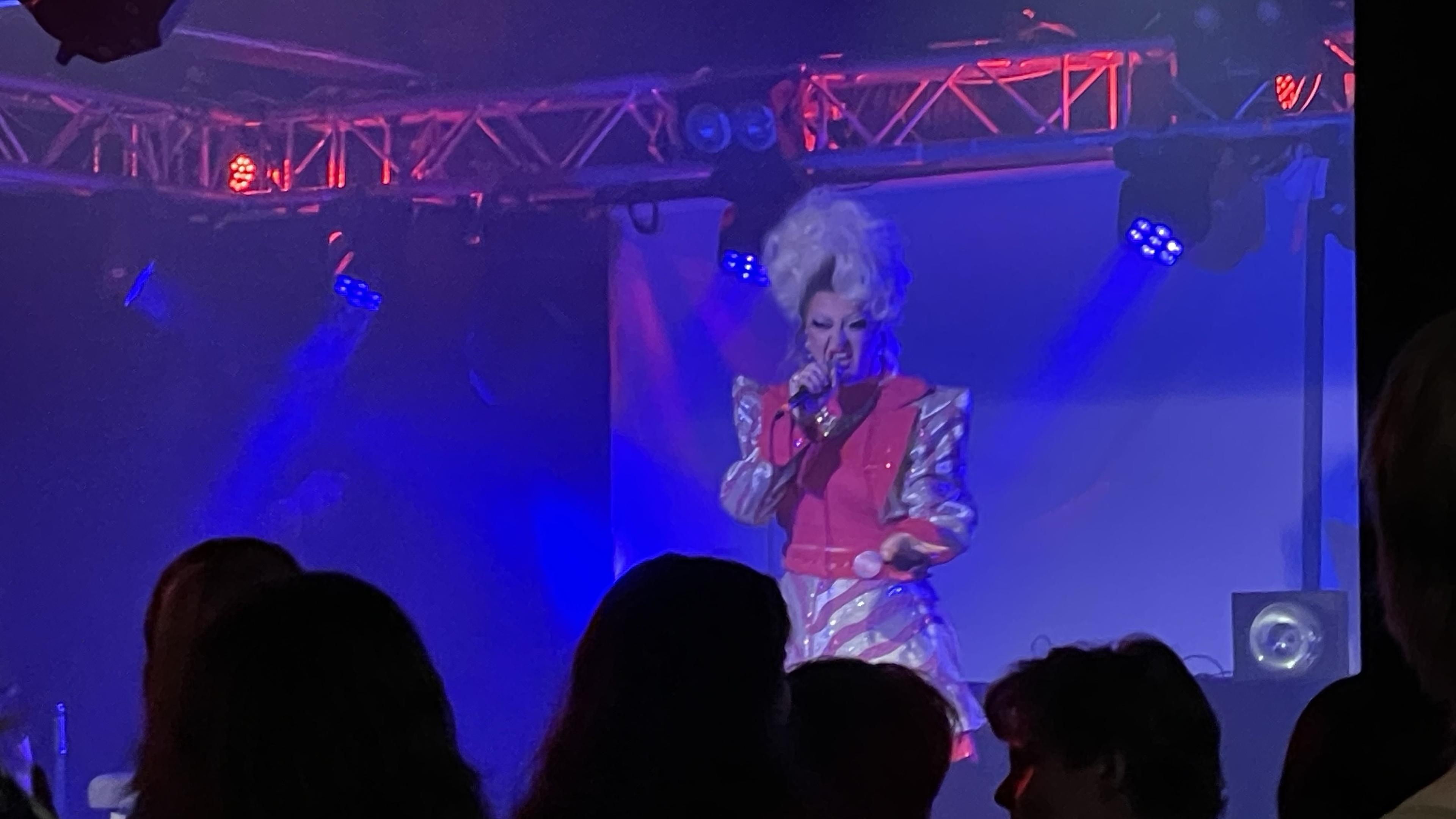 GeeGee Van De Slay lavalla. Drag-artisti punavalkeassa mekossa ja valkoisessa peruukissa.