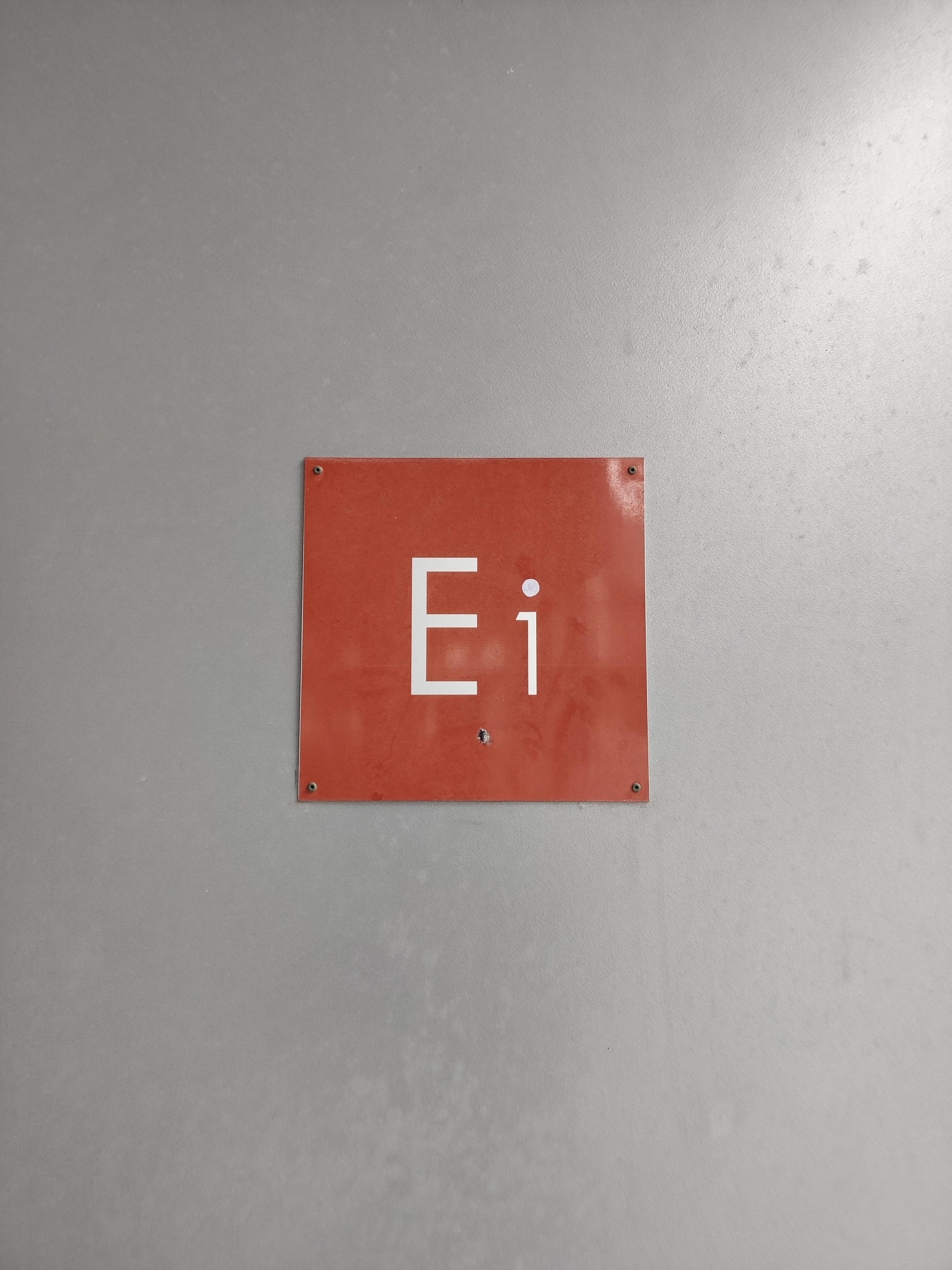 Punainen porraskäytävän opaste kyltti, jossa lukee valkoisella E1. Kyltissä numero on puolet pienempi kuin kirjain ja sen väkänen on vaakasuuntainen. Numeron päälle on piirretty valkoinen piste, jolloin näyttää, kuin kyltissä lukisi Ei.