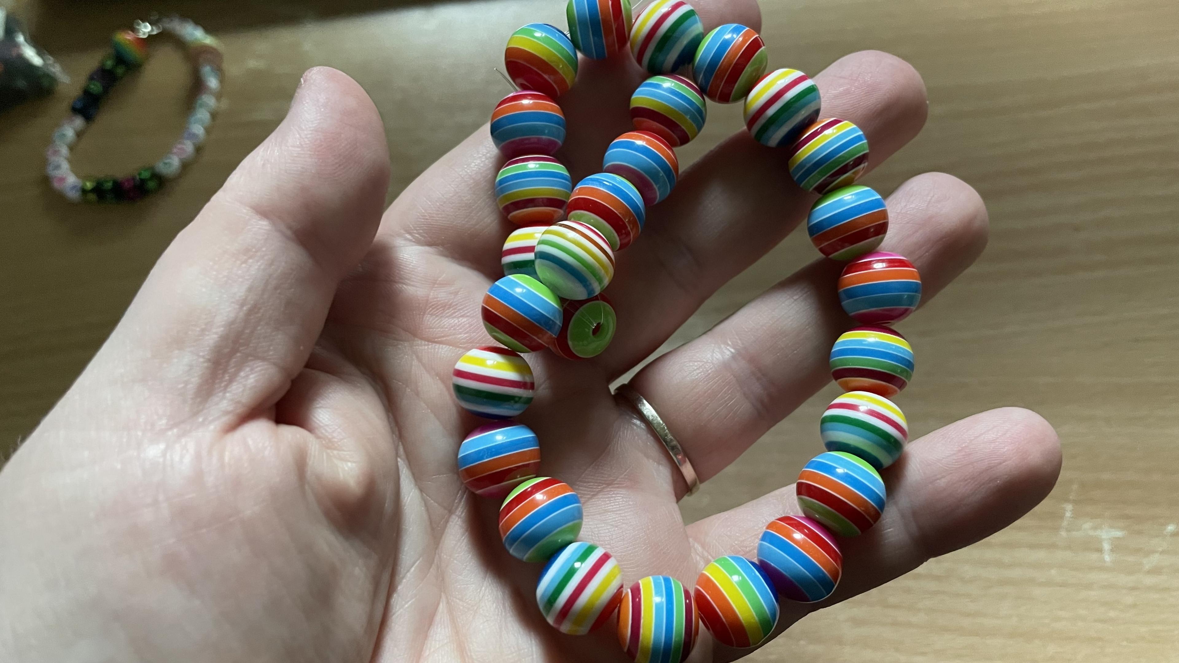 Round colorful plastic beads made into a bracelet on the palm of my left hand. Each bead has some sort of a rainbow combination of colors and they have been arranged red against red and blue against blue so they sort of have some sense in how the colors flow through the whole thing.